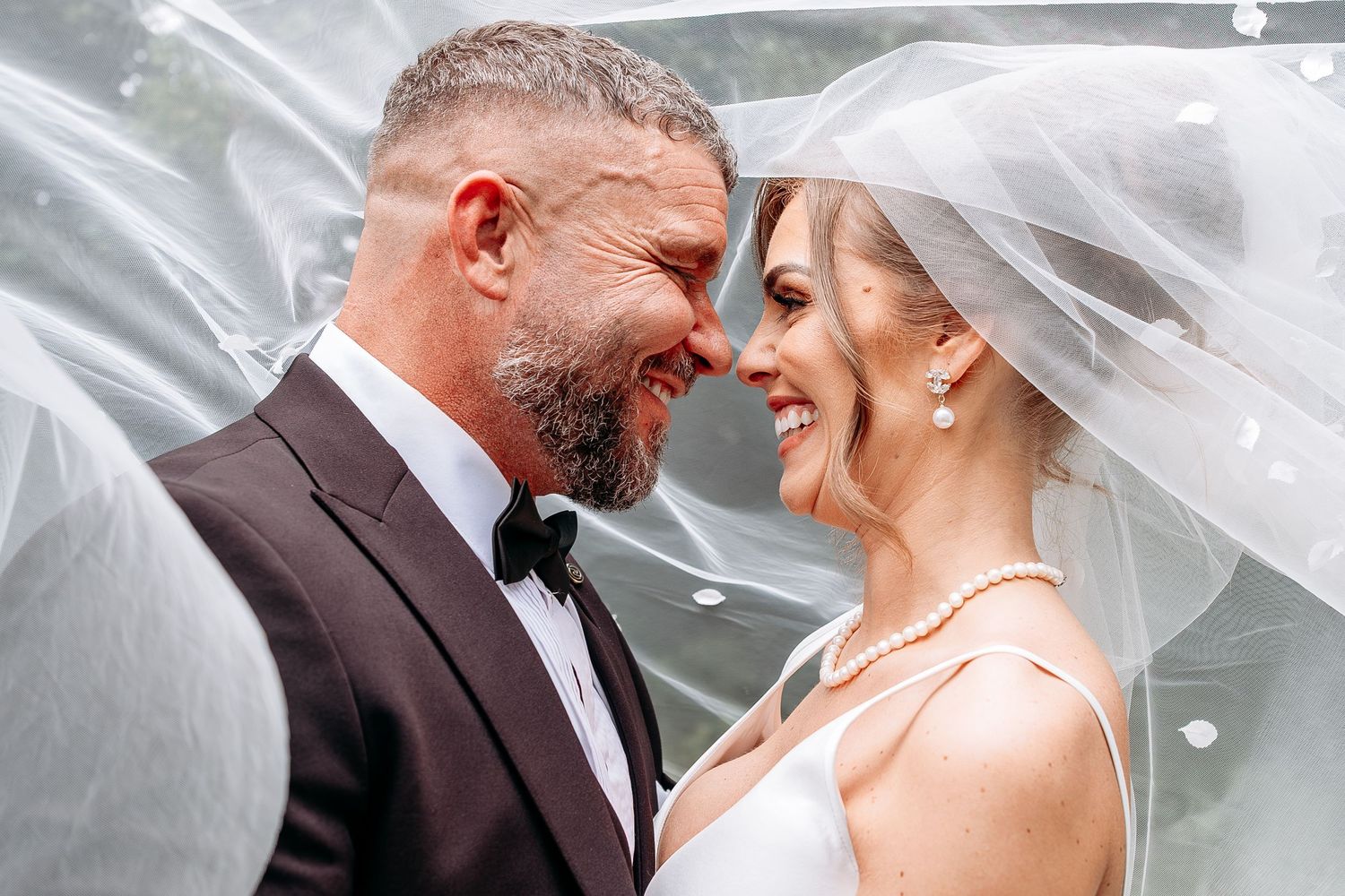 Happy newlyweds share a romantic moment under a flowing veil, groom in tuxedo and bride in white wedding dress.