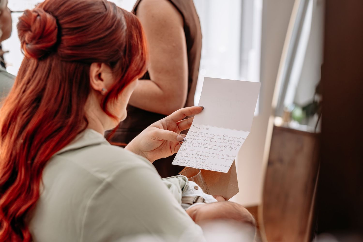 Red-haired woman reading a handwritten letter card, seated indoors near a window in casual attire.