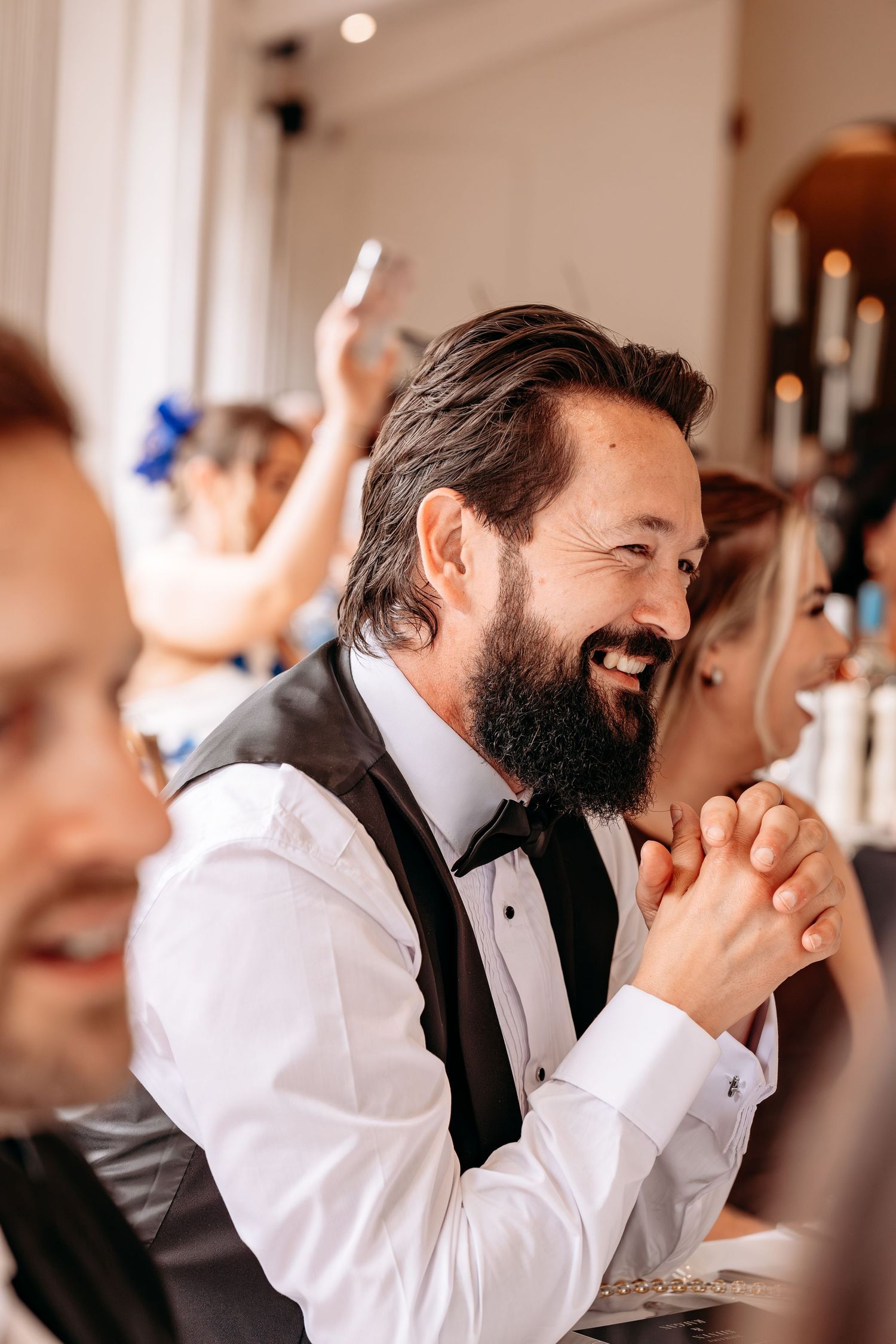 Bearded man in tuxedo vest smiling joyfully at a wedding reception, clapping hands together.