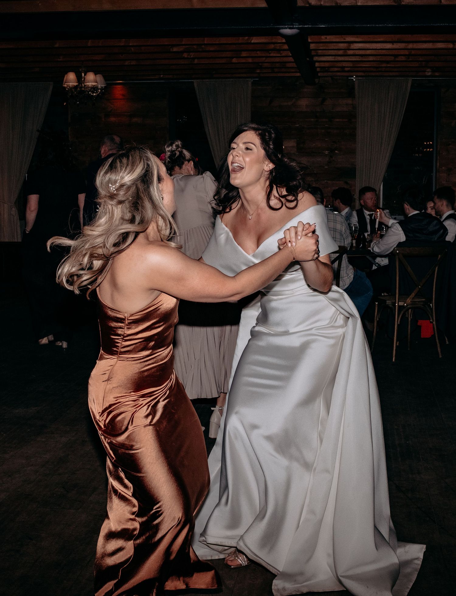 Bride in white gown dancing joyfully with bridesmaid in copper satin dress at elegant wedding reception.