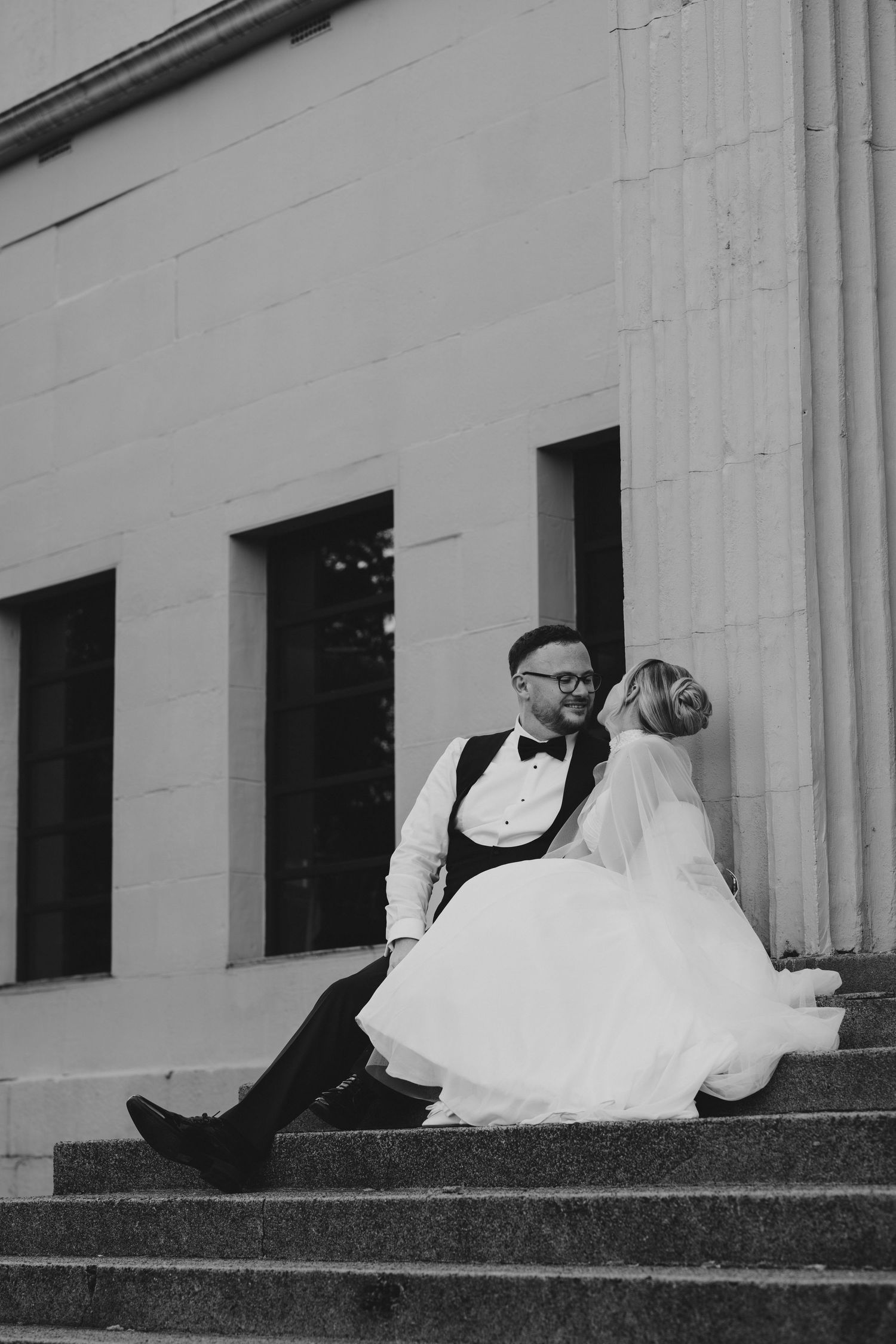 Black and white photo of bride and groom sitting on steps outside a grand building on their wedding day.