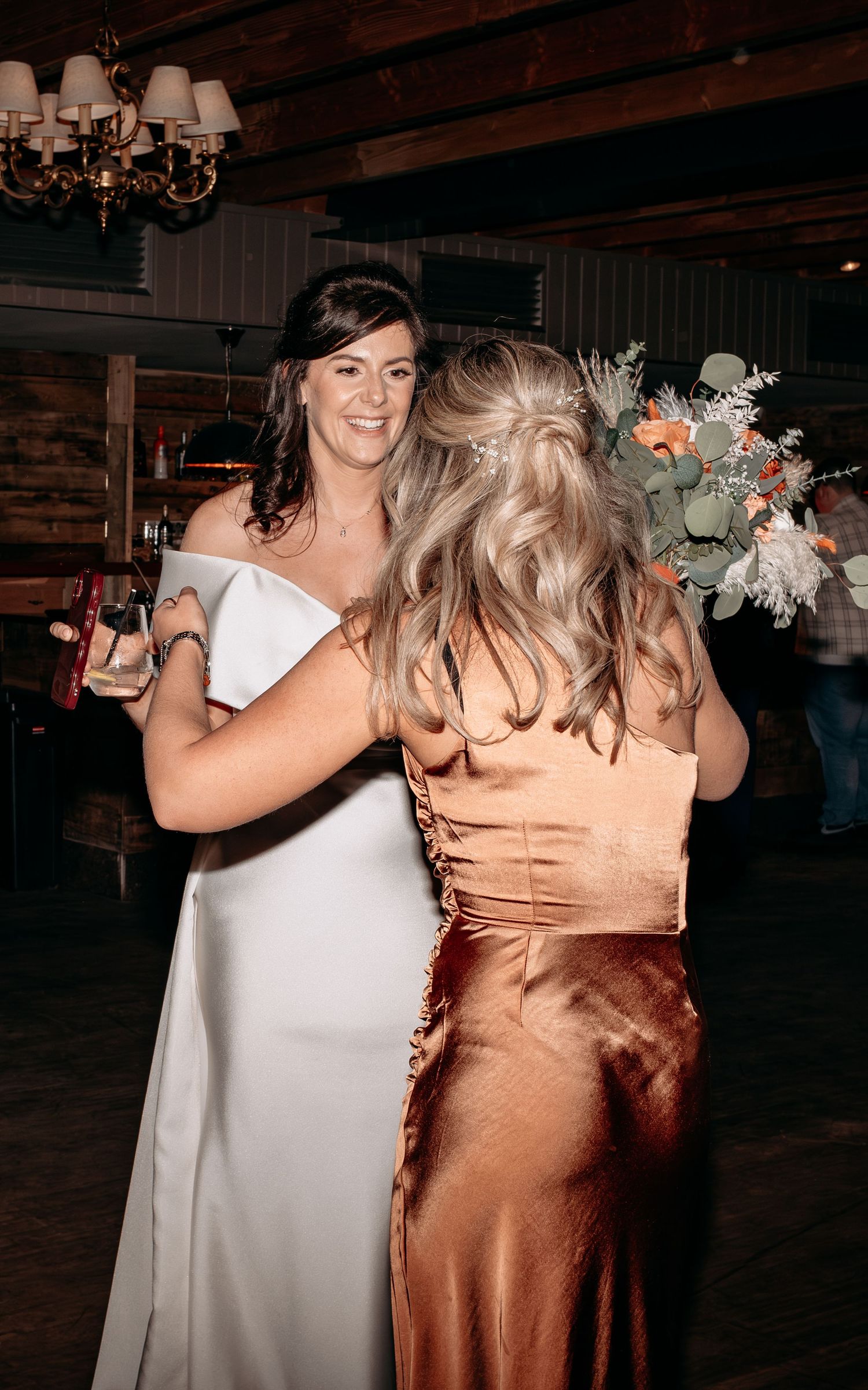 Bride in white gown hugging a guest in copper satin dress at a rustic wedding reception.