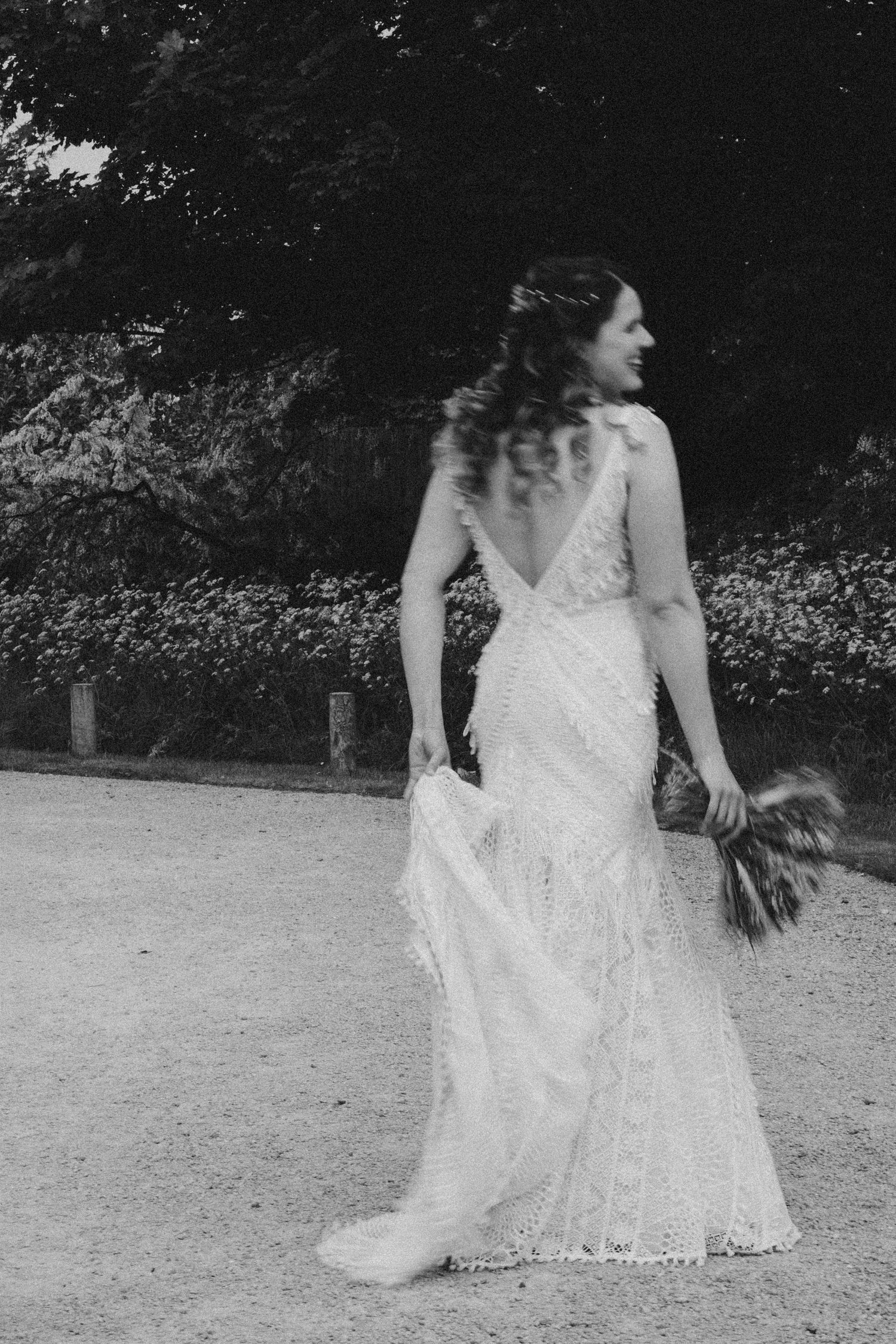 Black and white photo of a bride in a lace wedding gown holding a bouquet, walking outdoors.