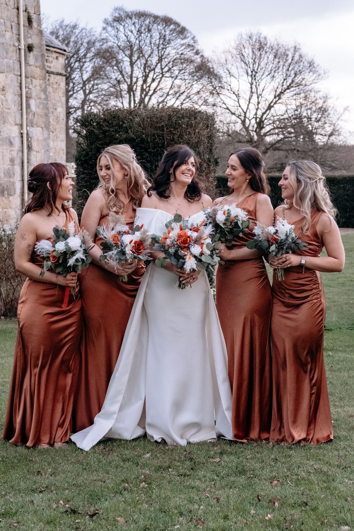Bride with four bridesmaids in burnt orange satin dresses holding bouquets outdoors.