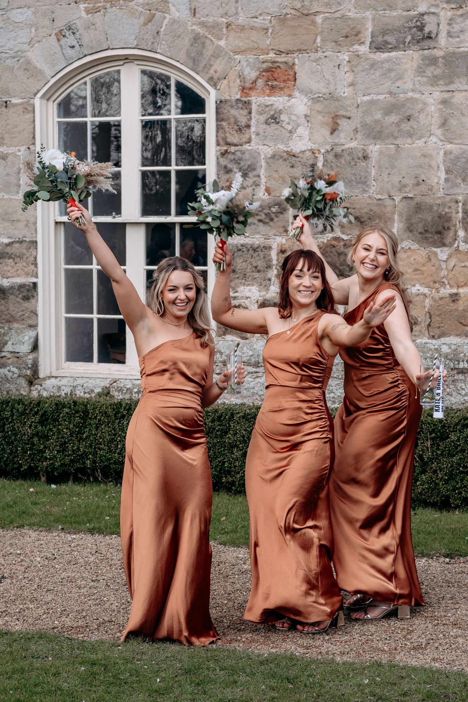 Three bridesmaids in rust satin gowns celebrate outdoors at a stone venue, raising bouquets and champagne glasses.