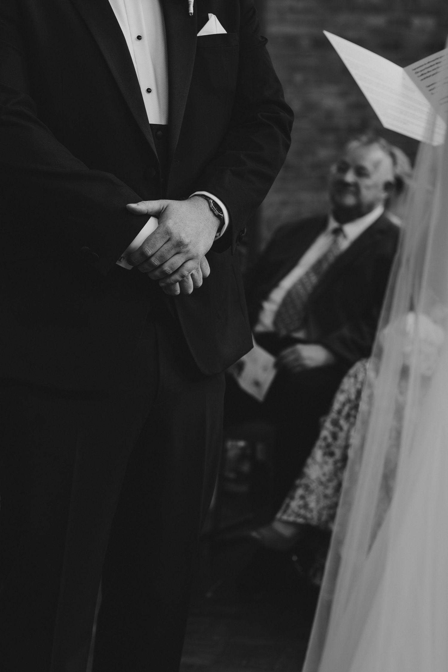 Black and white wedding photo of groom in tuxedo clasping hands at altar with seated guest in background.