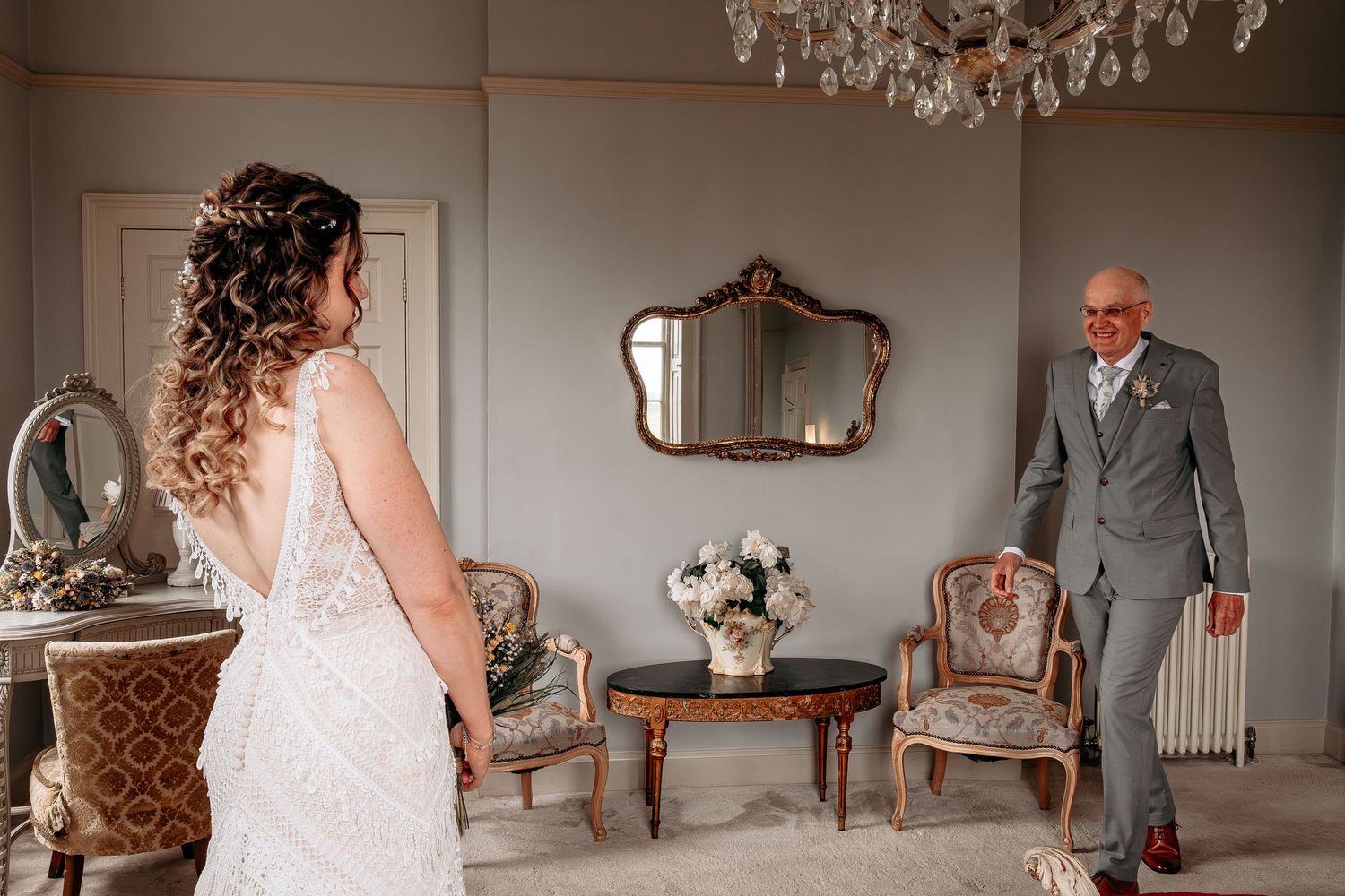 Bride in lace gown faces groom in gray suit during emotional first look in elegant vintage room with chandelier.