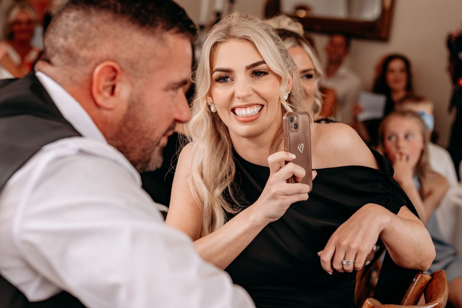 A smiling blonde woman in a black dress holds a phone at a social gathering with guests in the background.