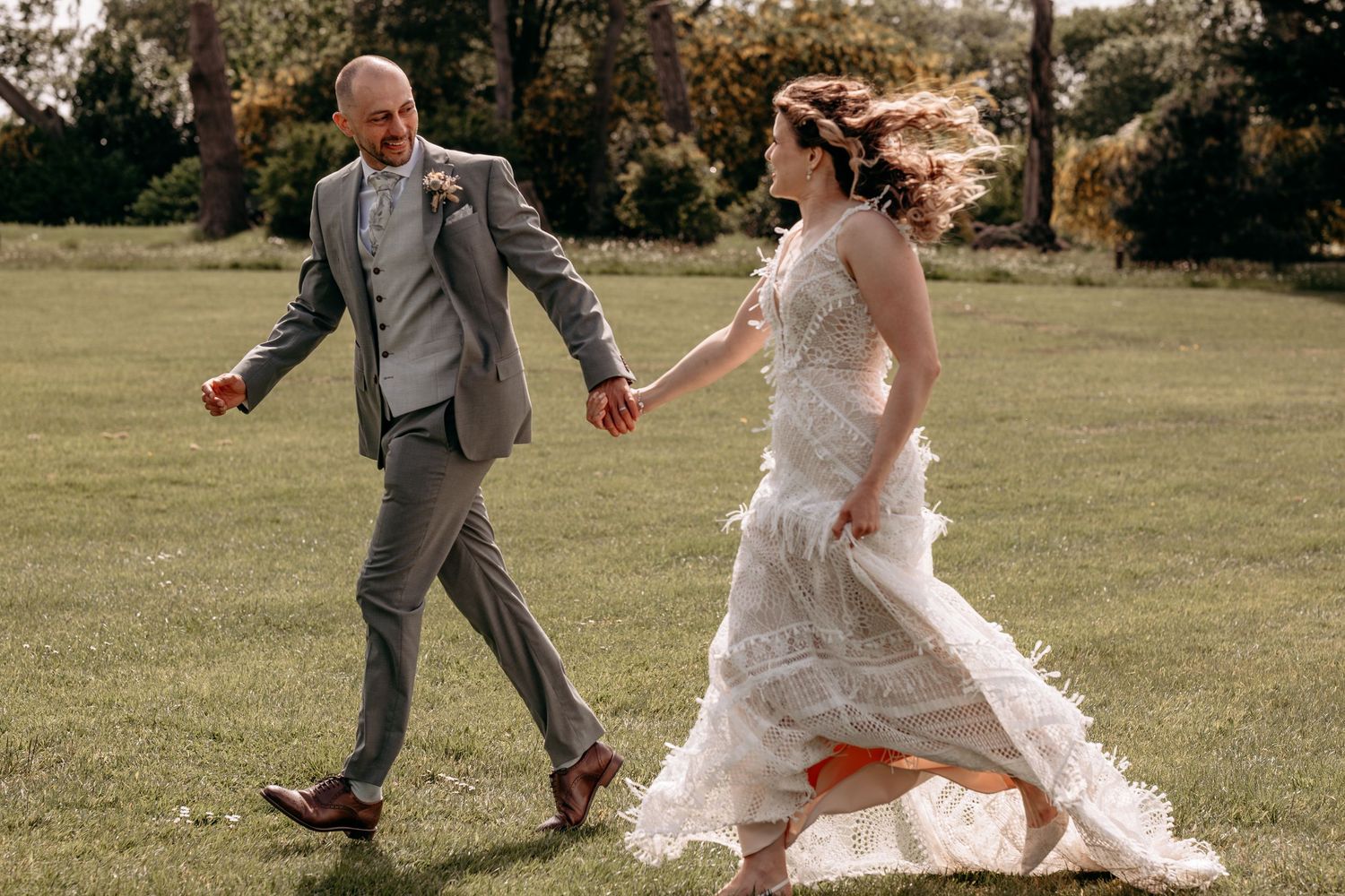 Bride and groom holding hands running across green lawn in wedding attire, white ruffled dress and gray suit.