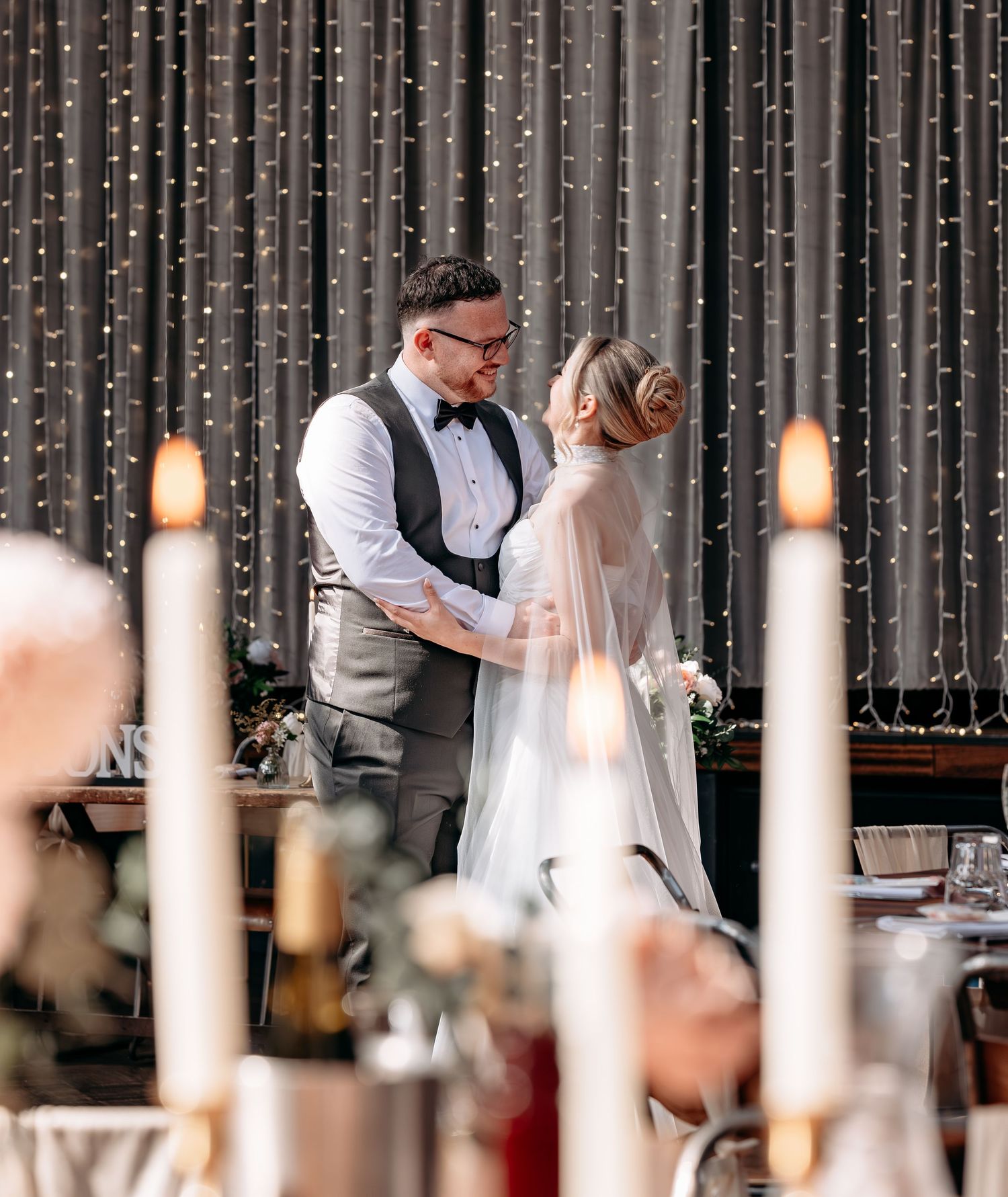 A bride and groom share a romantic moment at their wedding reception, framed by candlelight and twinkling fairy lights.