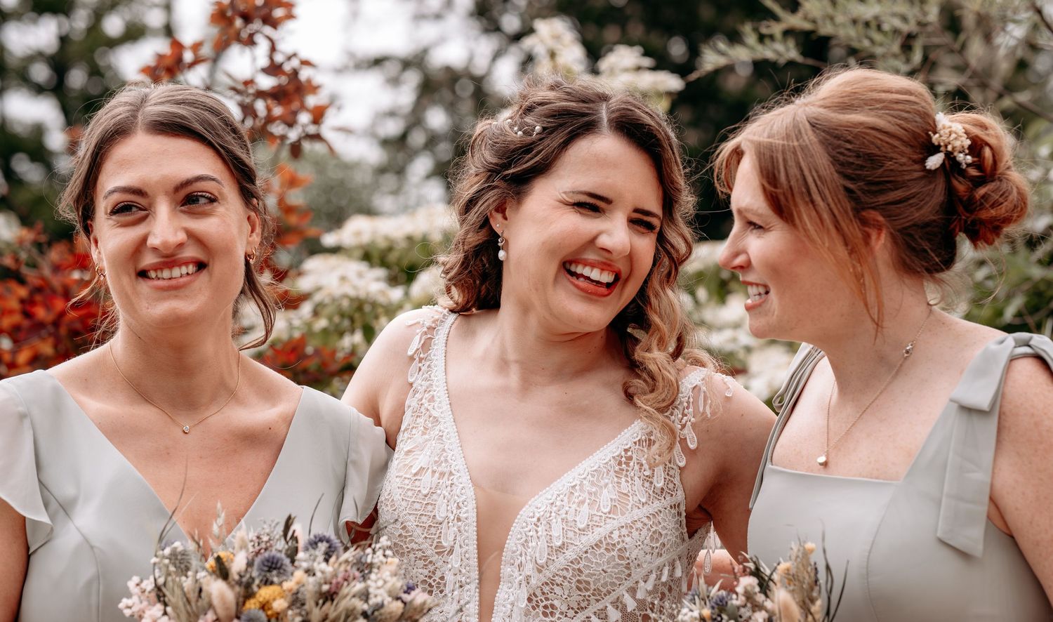 Bride laughing with two bridesmaids in sage dresses holding dried floral bouquets outdoors.