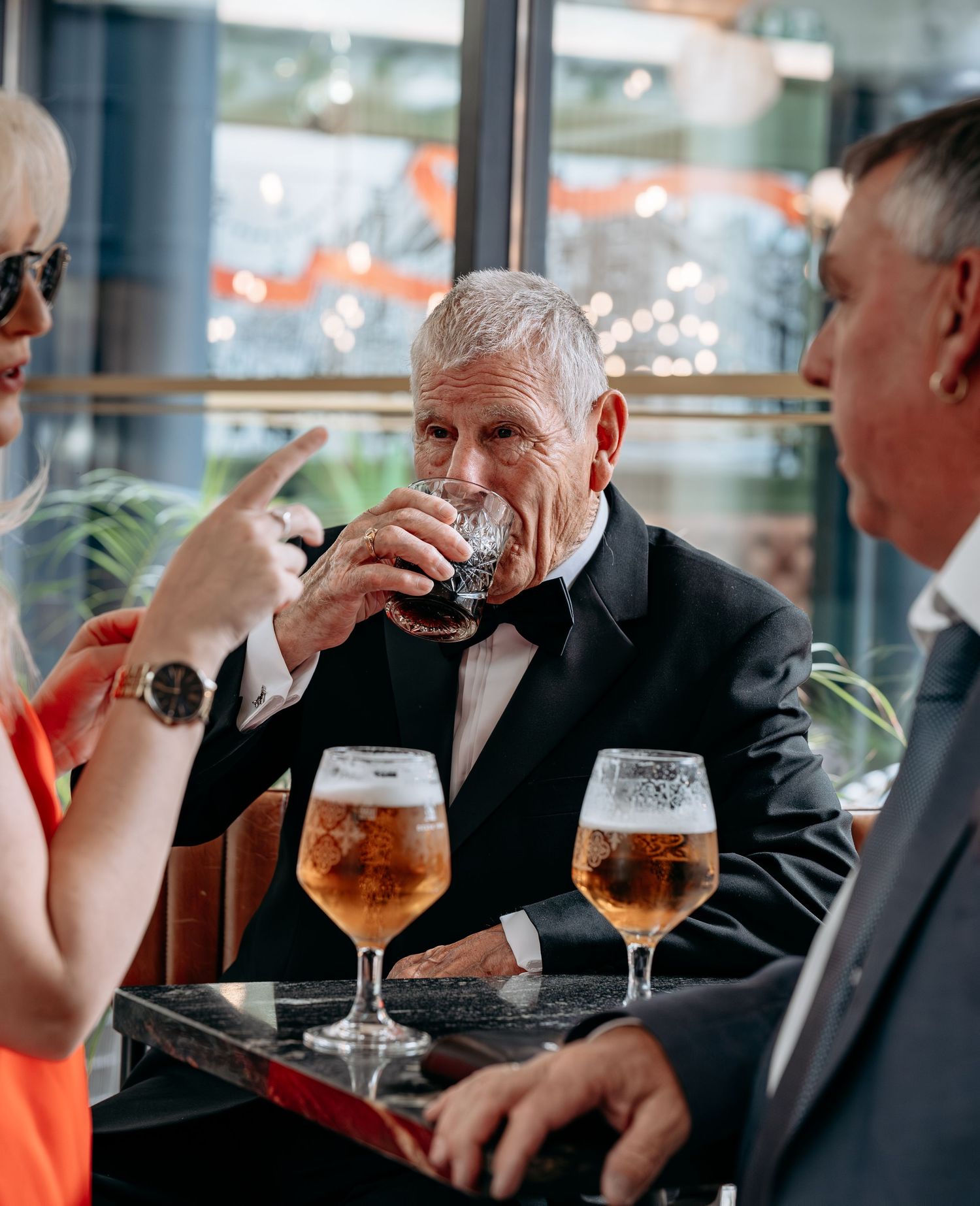 Elderly man in tuxedo drinking beer at table with wine glasses, socializing with guests at formal event.