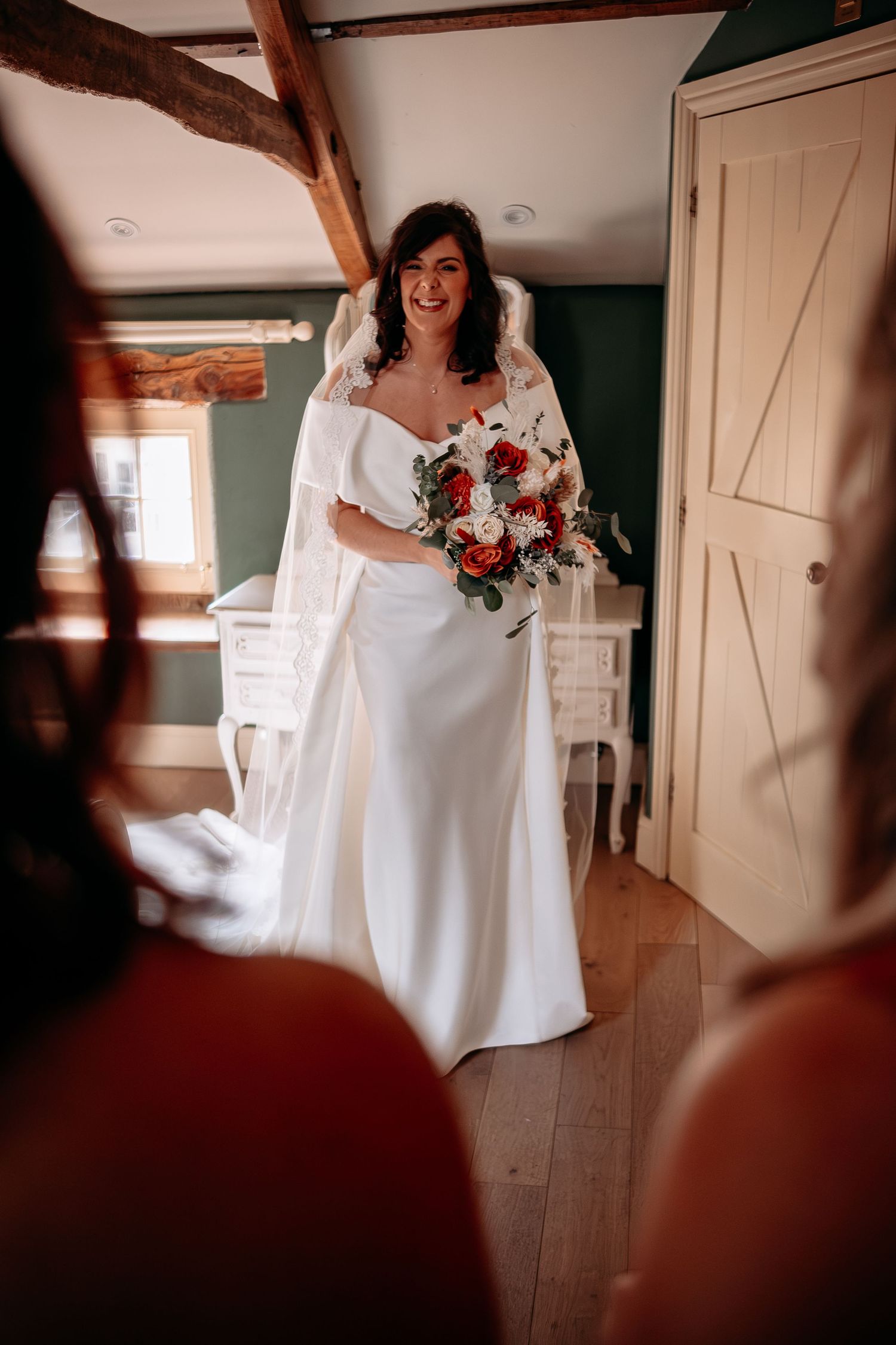 Smiling bride in white gown and lace cape holds red and white bouquet in a rustic wood-beamed room.