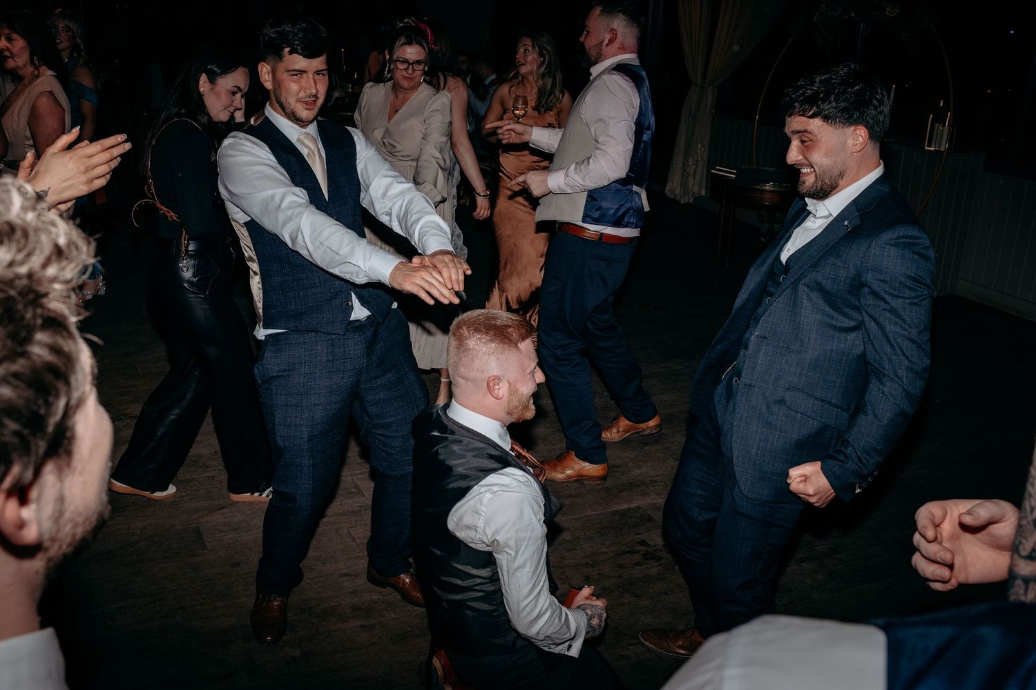 Groomsmen in navy suits dancing energetically at a wedding reception party on the dance floor.