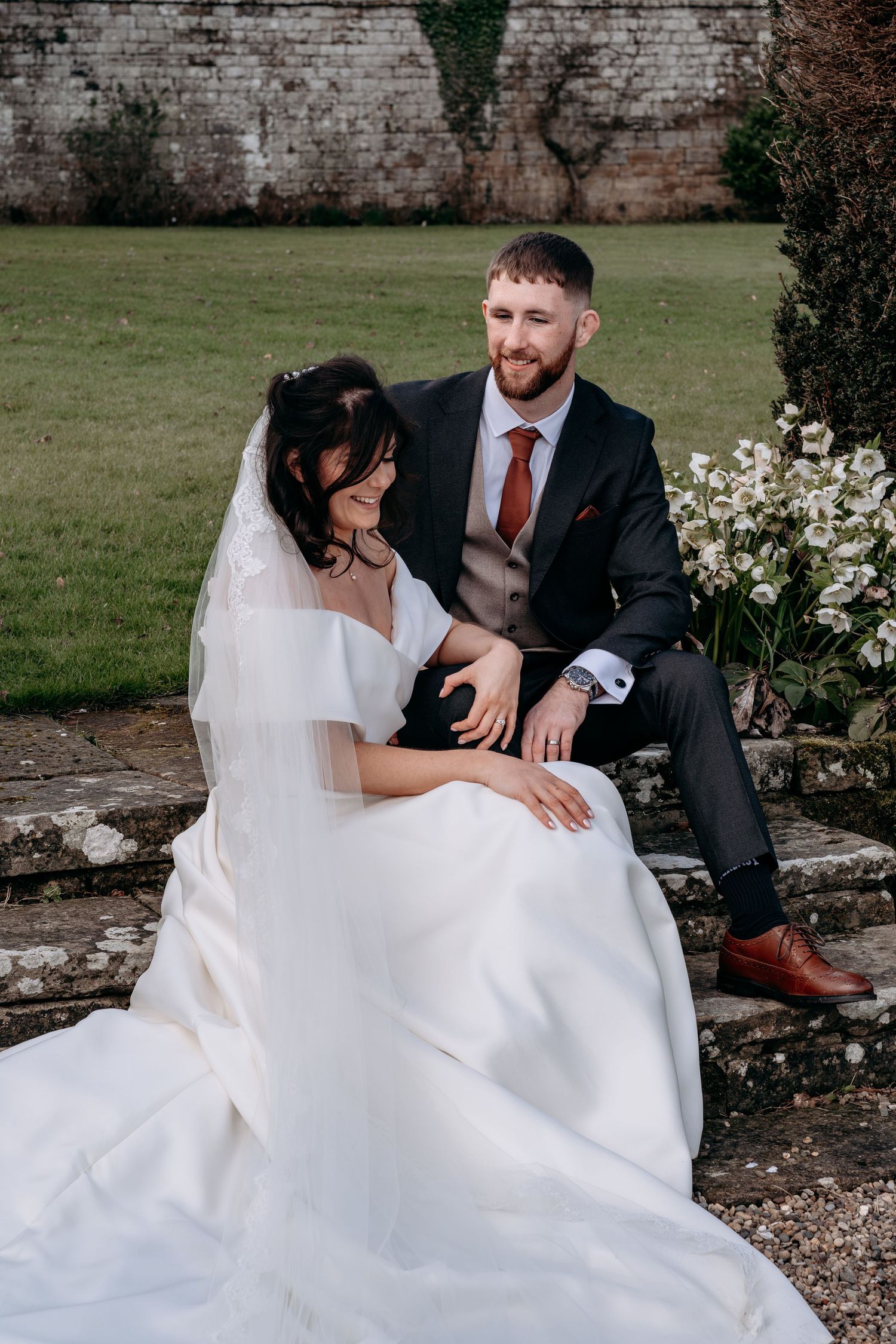 Bride and groom sitting on stone steps in a garden, bride in white gown with veil, groom in dark suit with burgundy tie.
