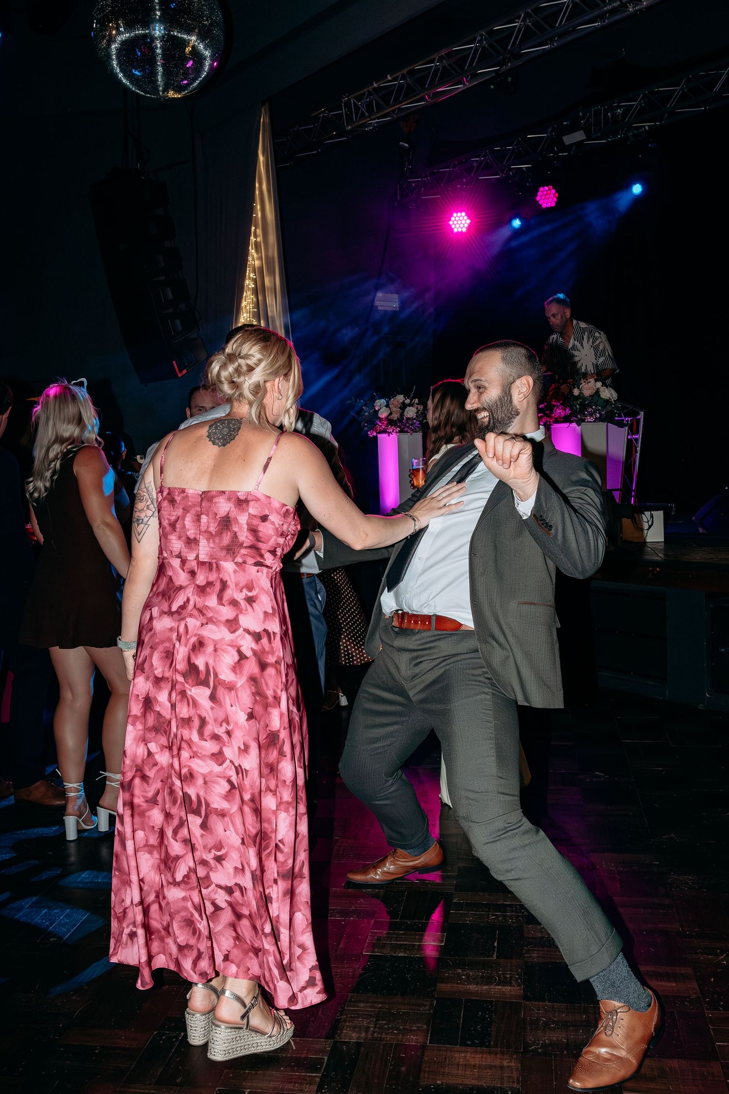 Couple dancing energetically at a wedding reception with pink stage lights and a disco ball overhead.