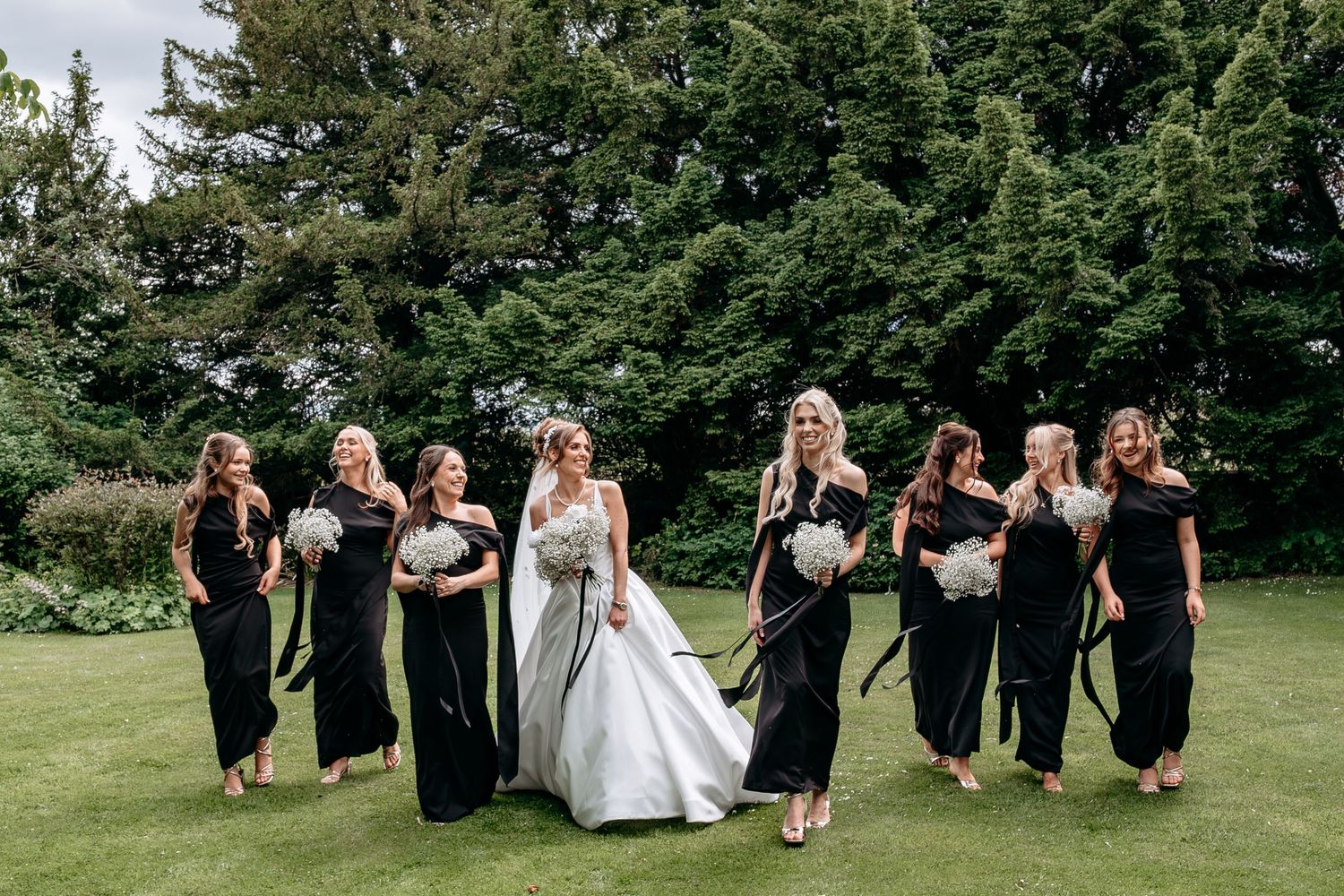Bride in white gown walks with bridesmaids in black dresses holding bouquets on green lawn with trees.