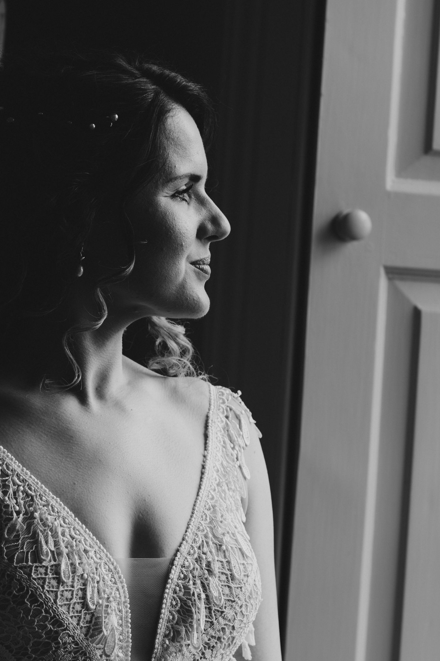 Black and white portrait of a smiling bride in lace dress gazing out a window on her wedding day.