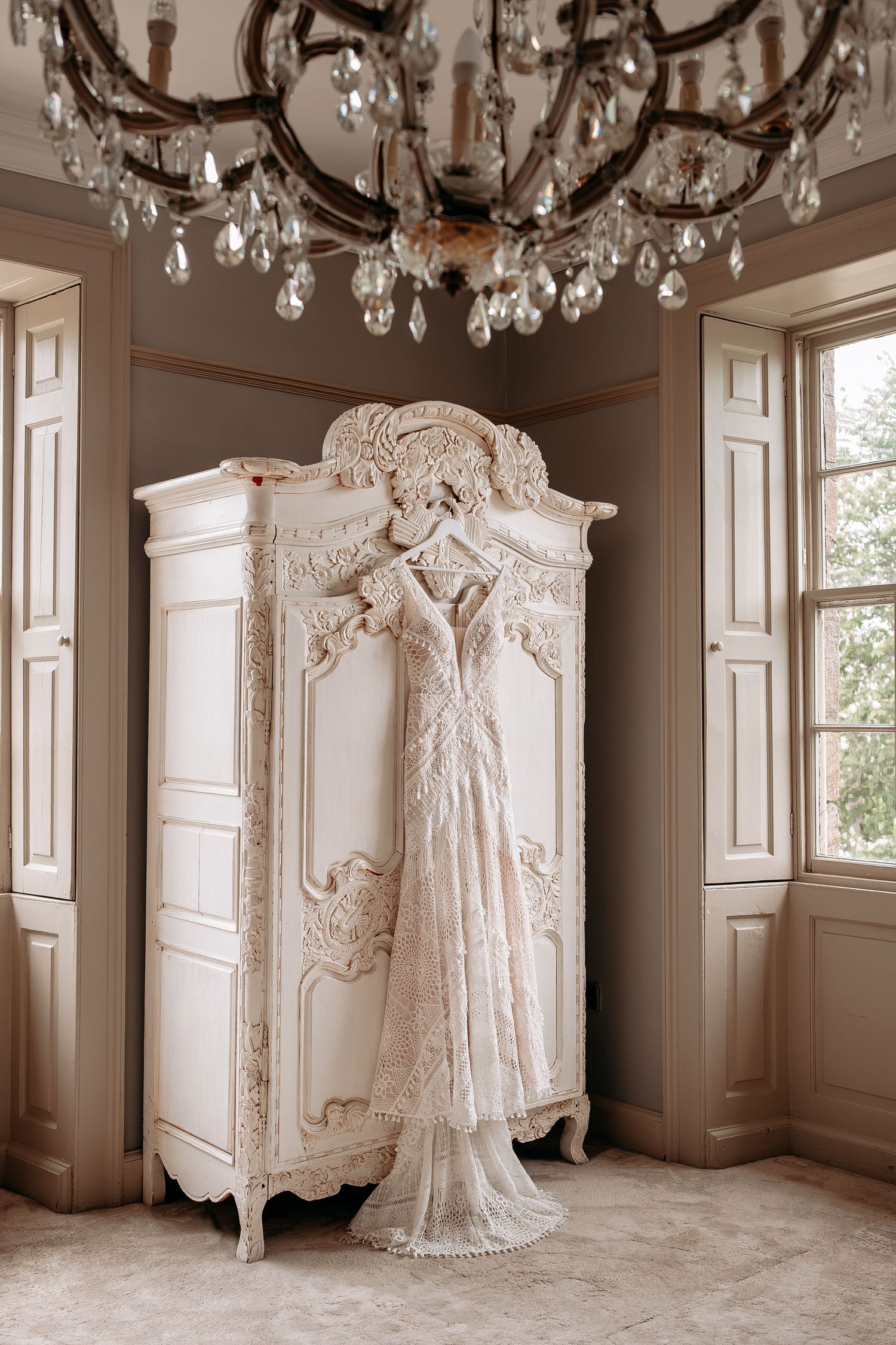 Elegant lace wedding dress hanging on ornate white armoire beneath crystal chandelier in a vintage-styled bridal suite.