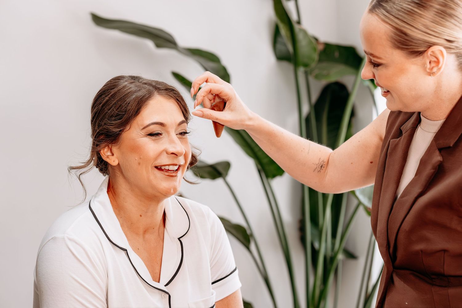 A makeup artist applying cosmetics to a smiling woman in a white top, with a green plant in the background.