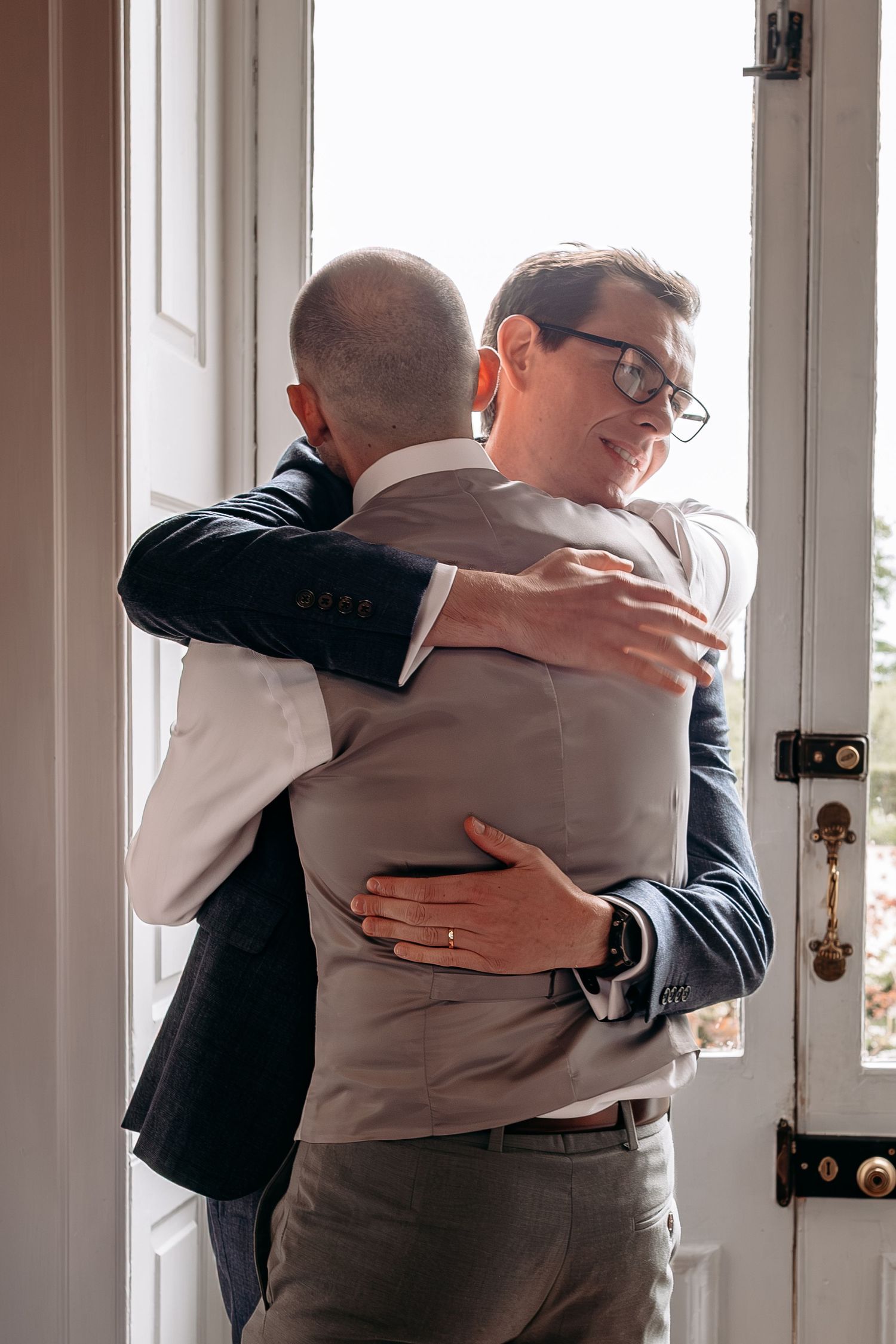 Two men in suits share a warm hug near a bright doorway, one wearing glasses and a gray vest.