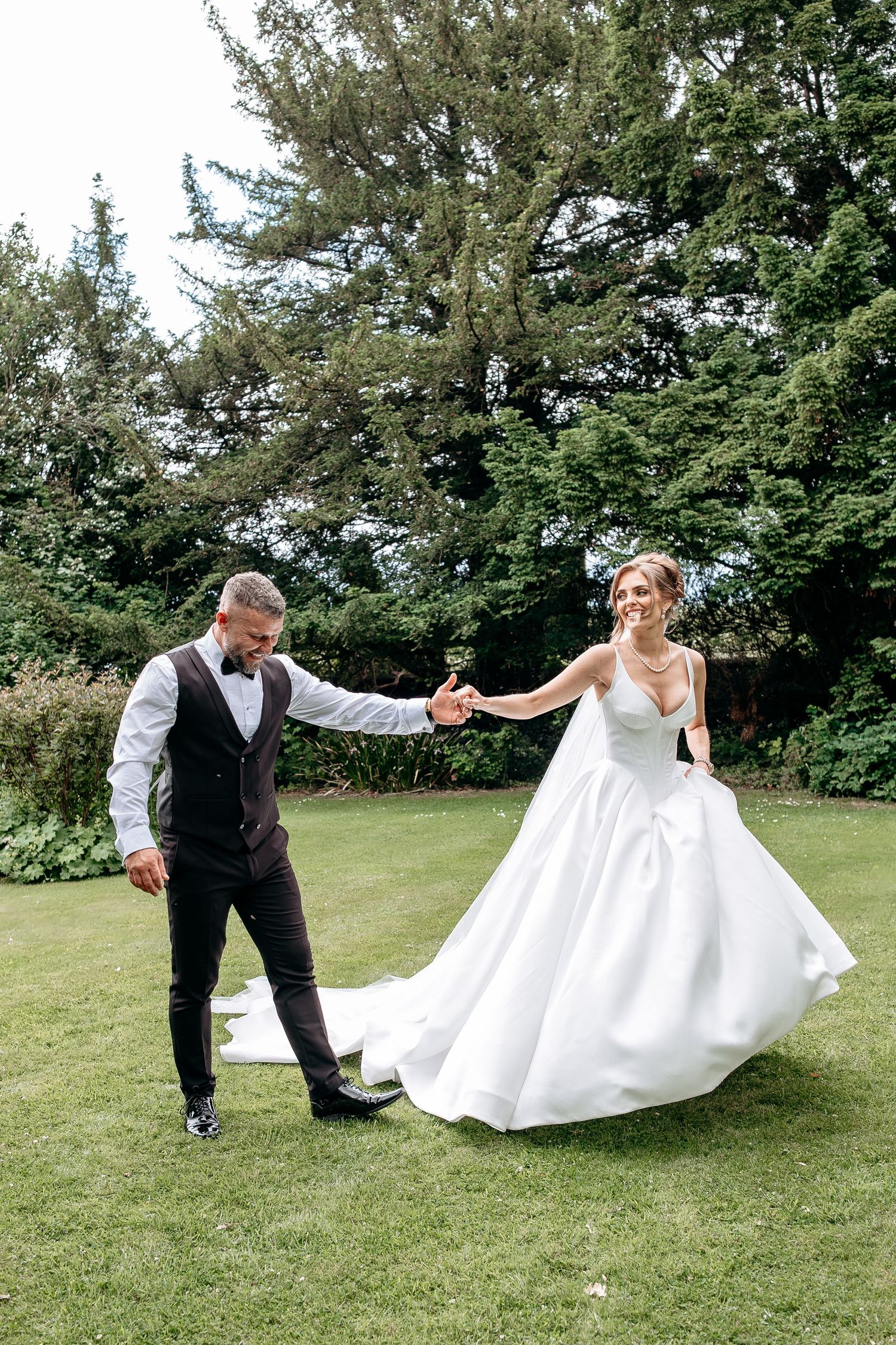 Bride in white ball gown and groom in dark suit dance joyfully on green lawn surrounded by tall trees.