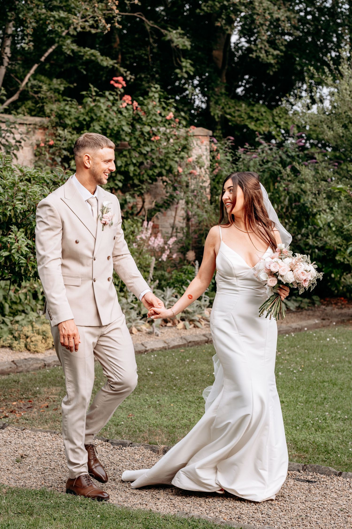 Bride and groom walk hand in hand through a lush garden, bride in white gown holding bouquet, groom in beige suit.