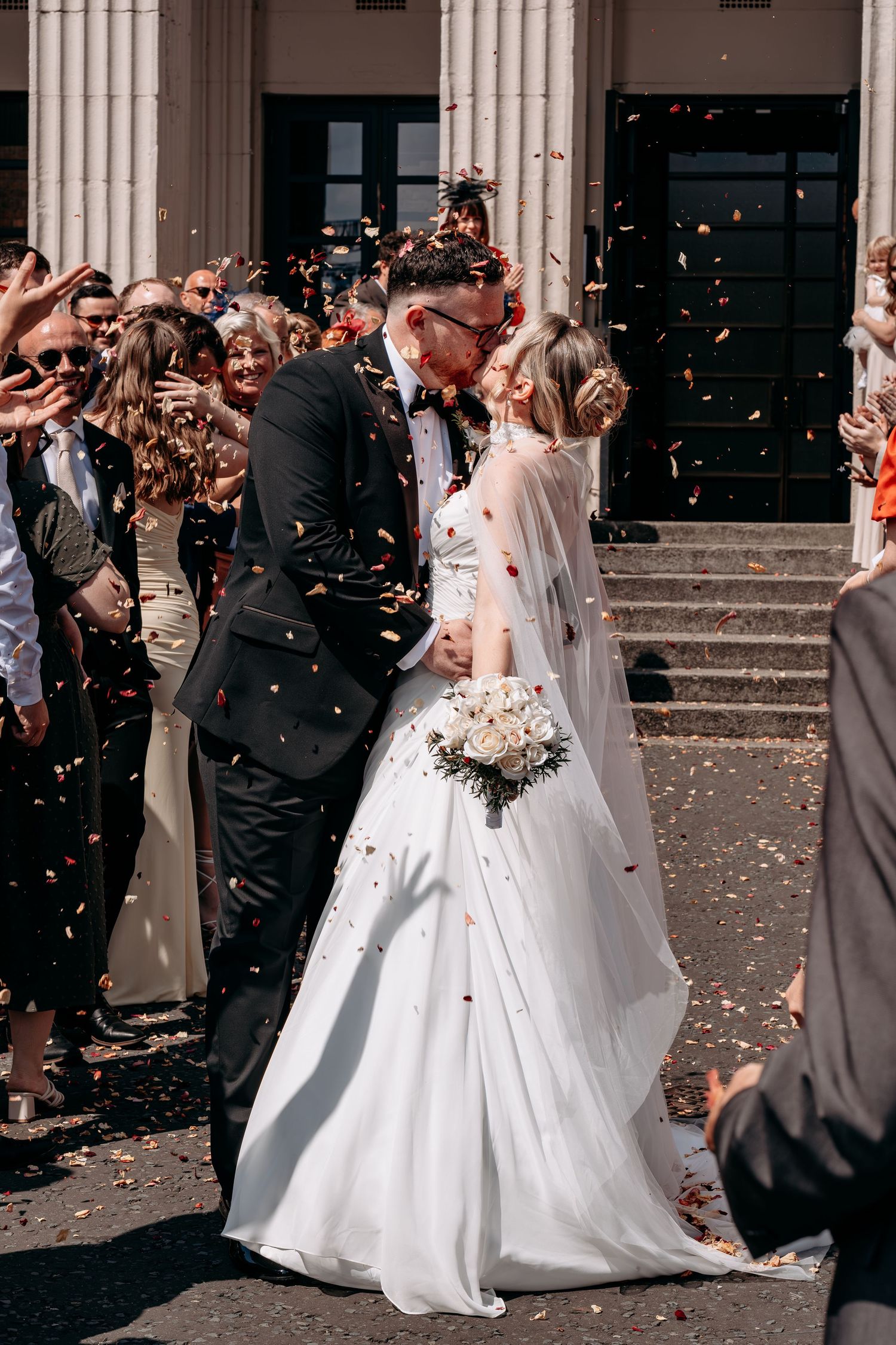 Bride and groom share a kiss outside church as guests throw colorful confetti during their wedding celebration.