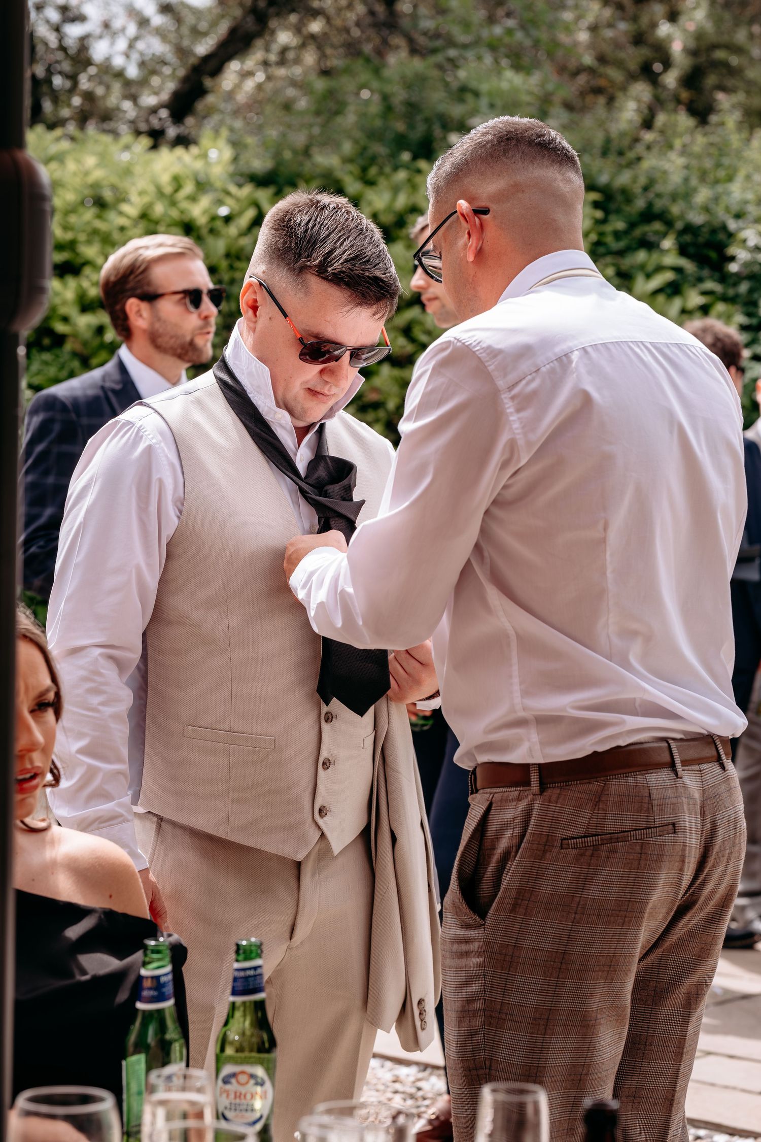 Groomsmen adjusting suit jacket outdoors at a wedding reception on a sunny day.
