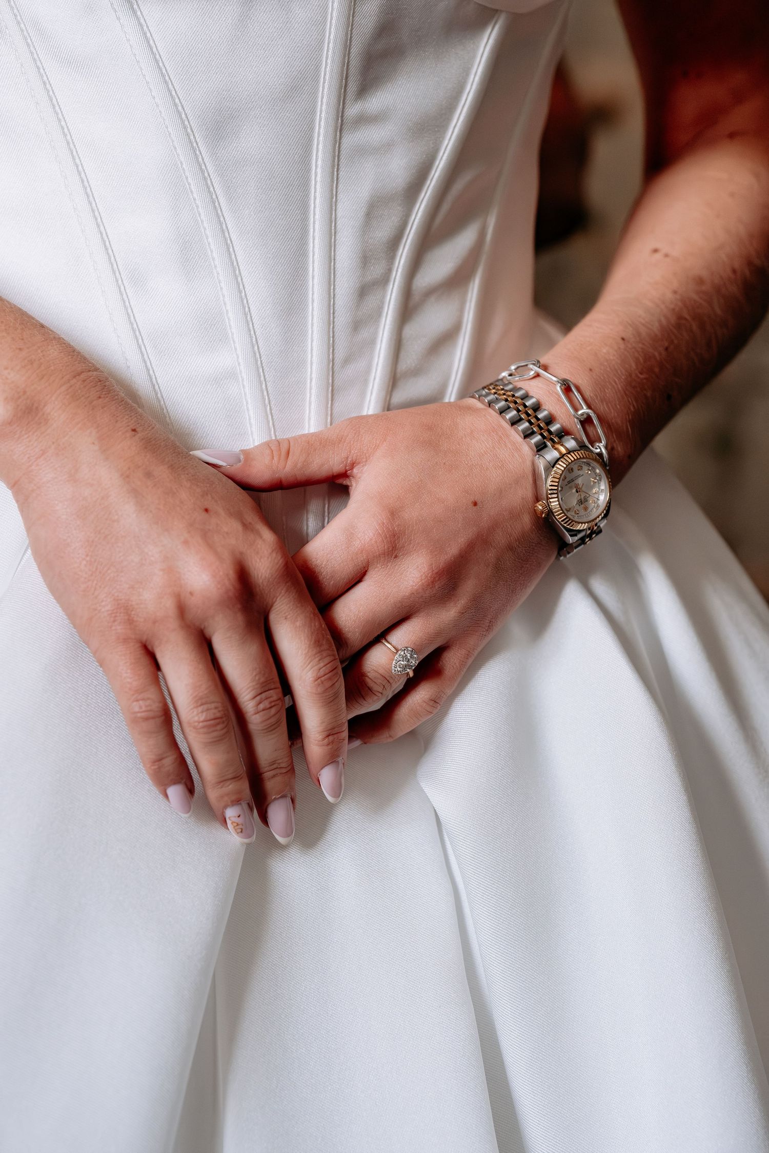 Close-up of bride's hands in white wedding dress, wearing diamond ring and two-tone Rolex watch.