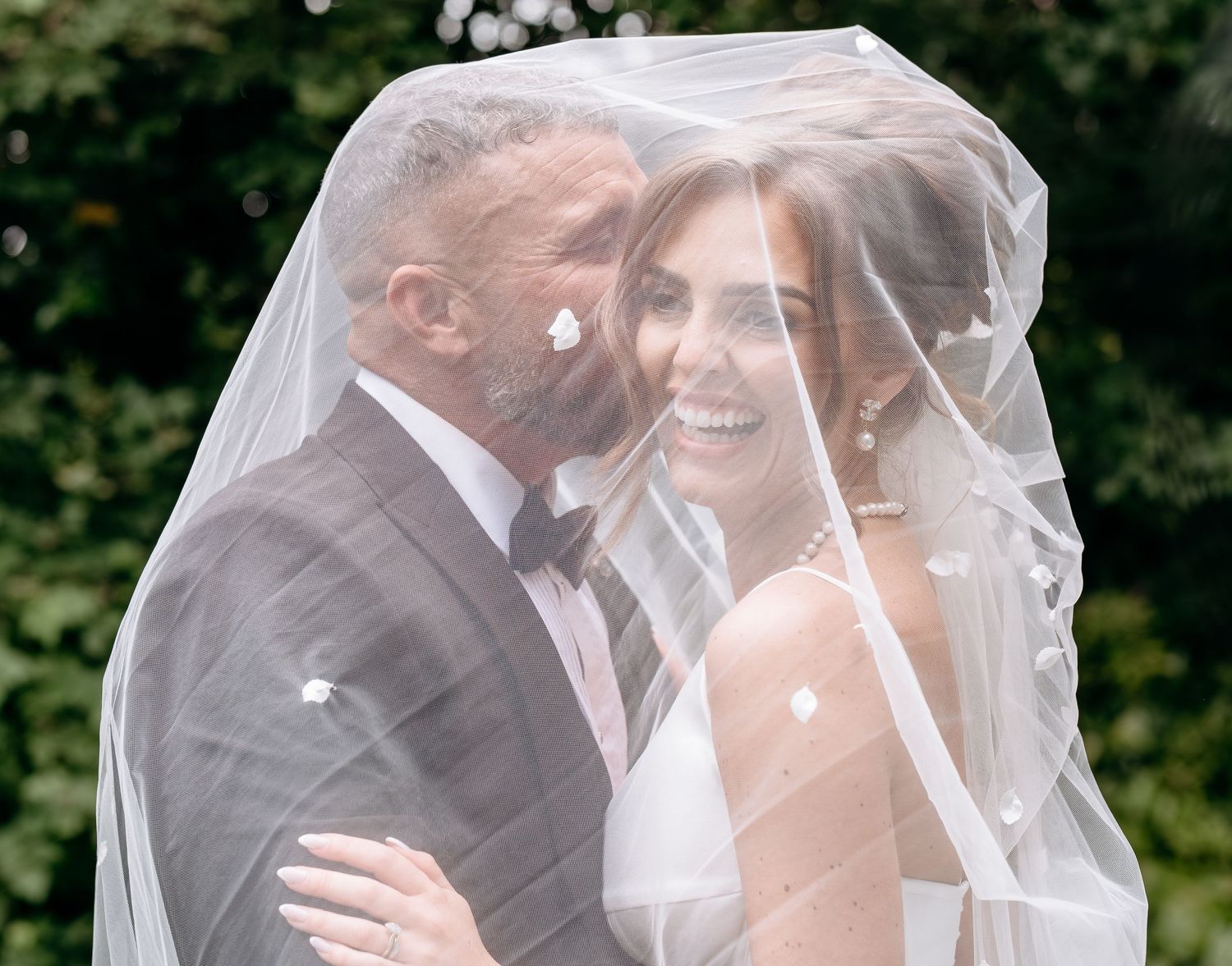Bride and groom share a romantic kiss under a sheer wedding veil outdoors, surrounded by lush greenery.