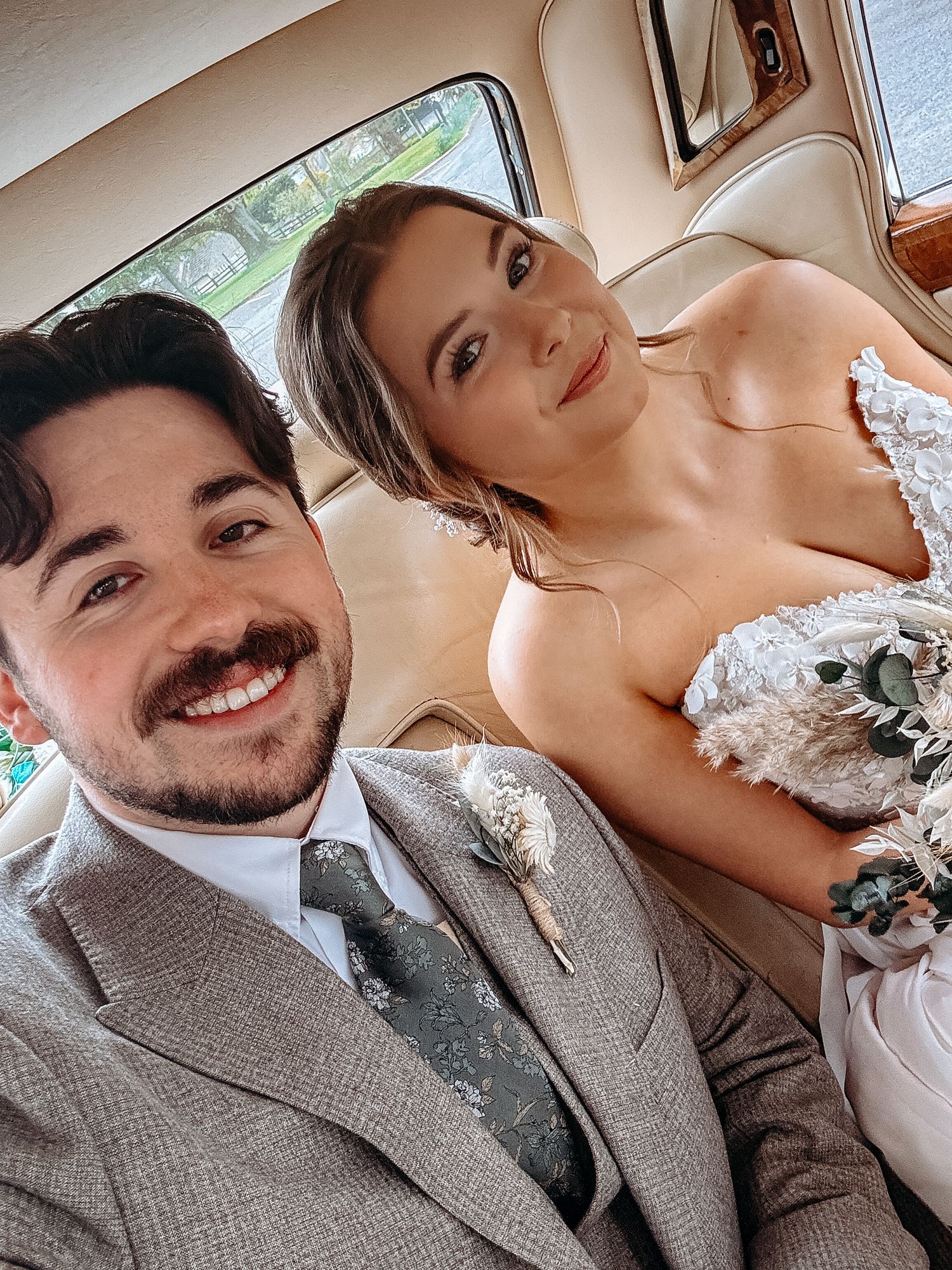 A smiling groom in a gray suit with boutonniere takes a selfie with his bride in a white gown inside a wedding car.