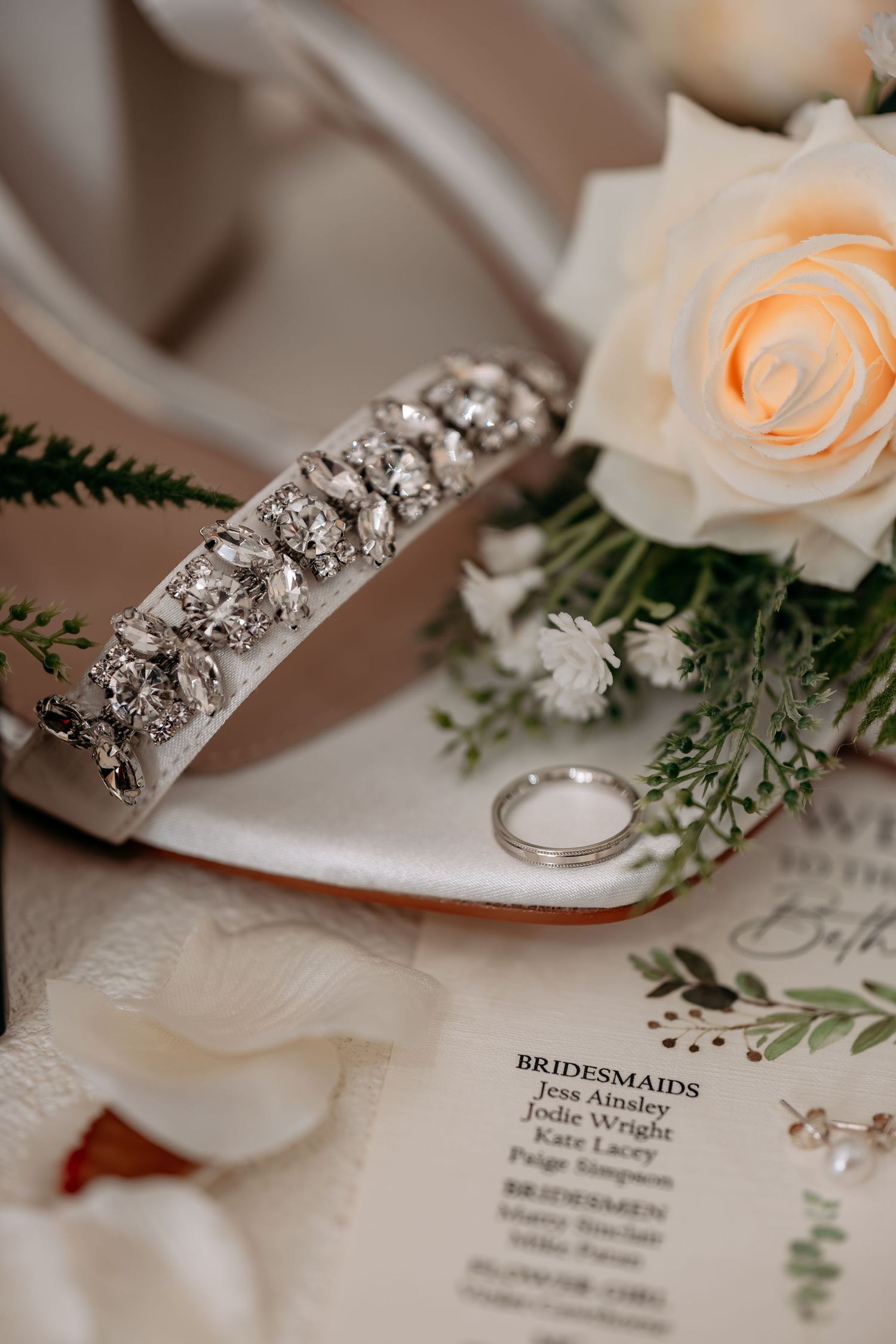 Elegant bridal flat lay featuring crystal-embellished wedding shoes, white roses, rings, and a bridesmaid card.