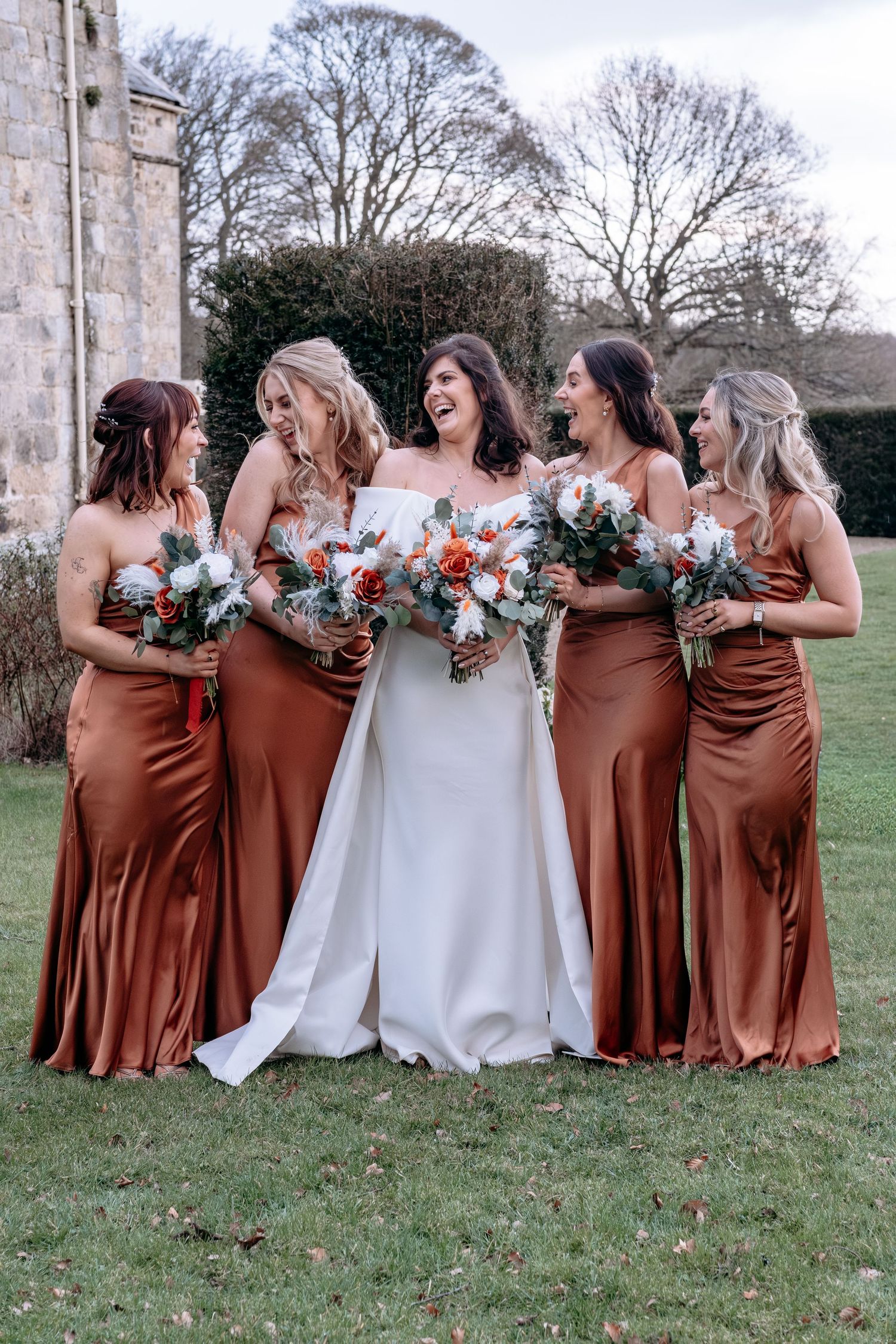 Bride in white gown laughs with four bridesmaids in rust satin dresses holding bouquets outdoors.