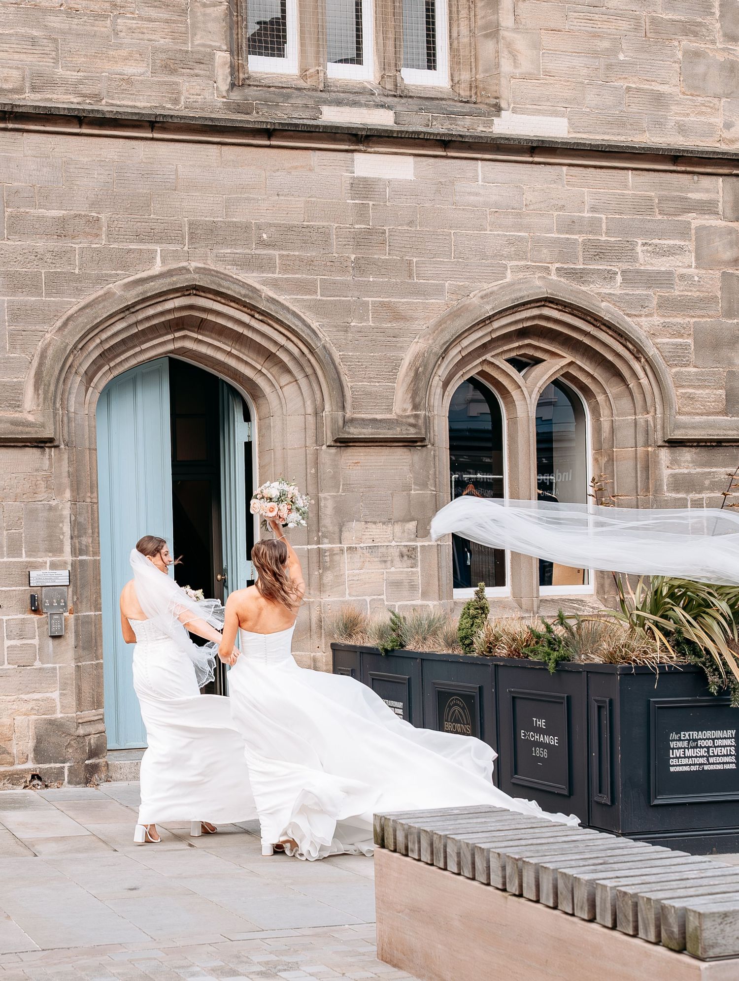 Two brides in white gowns with flowing veils approach a Gothic stone church entrance on their wedding day.