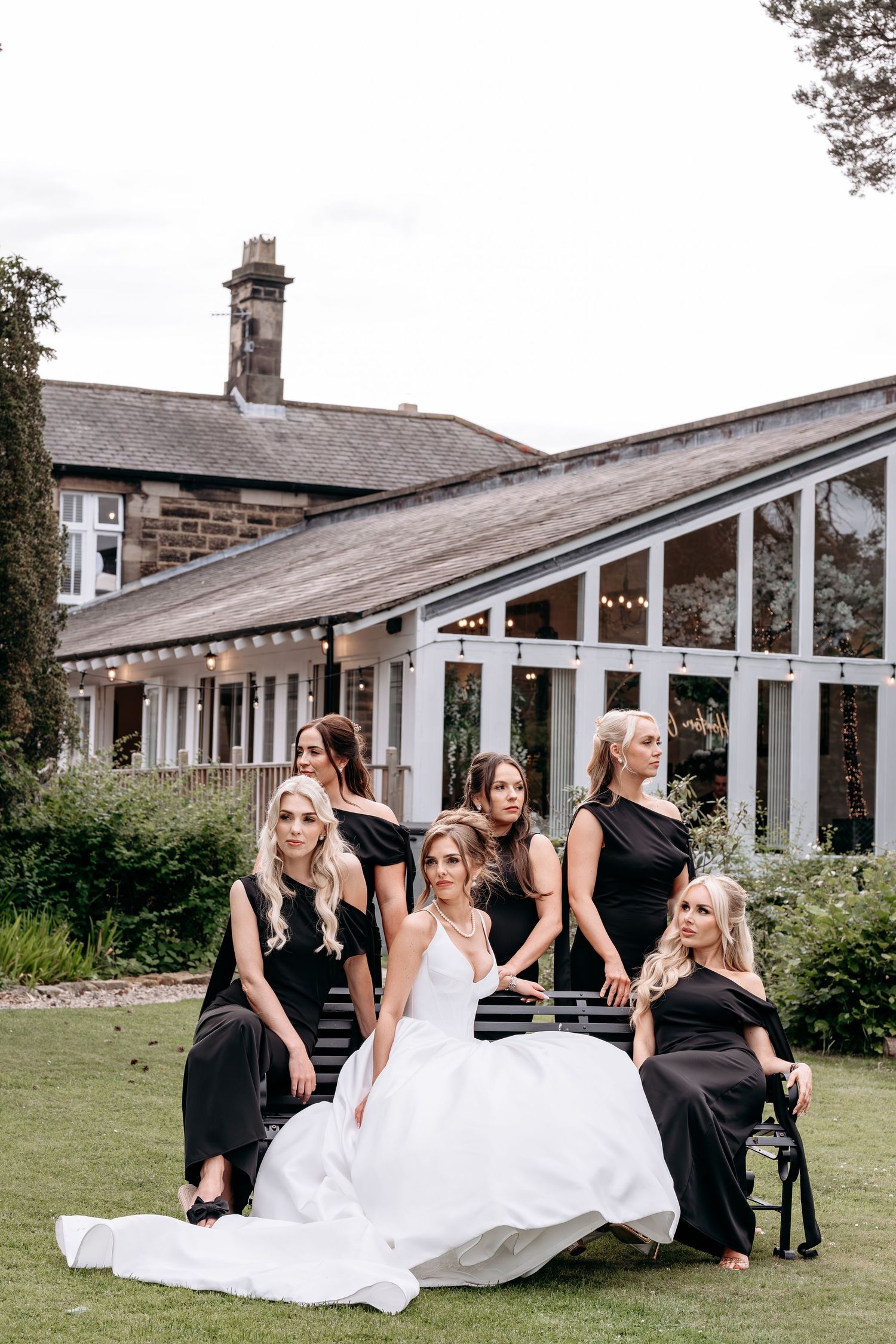 Bride in white gown poses with bridesmaids in black dresses on garden bench outside elegant stone venue.