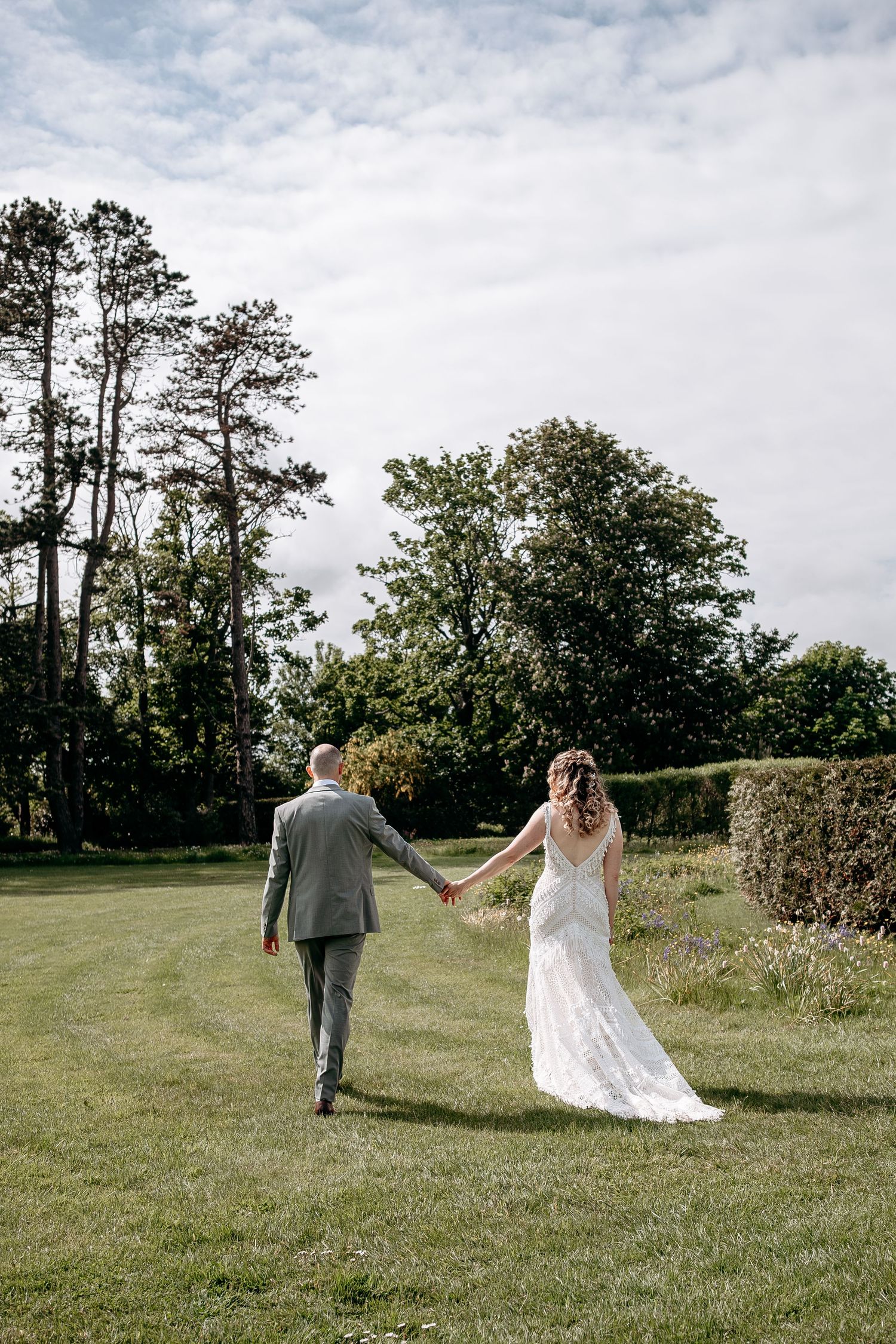 A bride and groom walk hand in hand across a lush green lawn surrounded by trees on their wedding day.