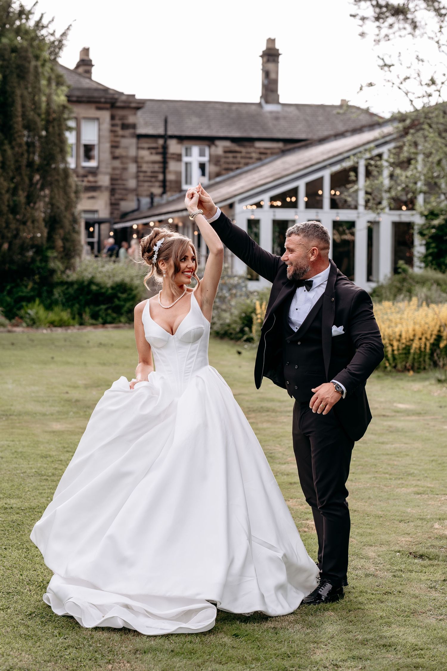 A bride and groom dancing joyfully on a green lawn outside a stone manor house on their wedding day.