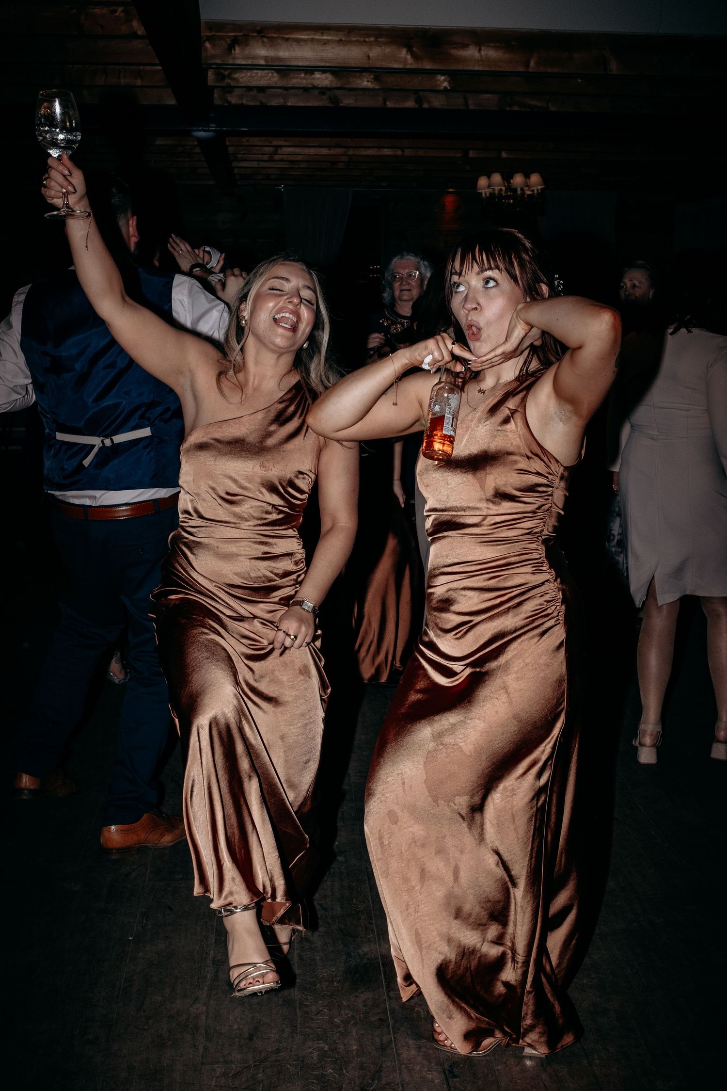 Two bridesmaids in satin bronze gowns dancing joyfully at a wedding reception, holding drinks.