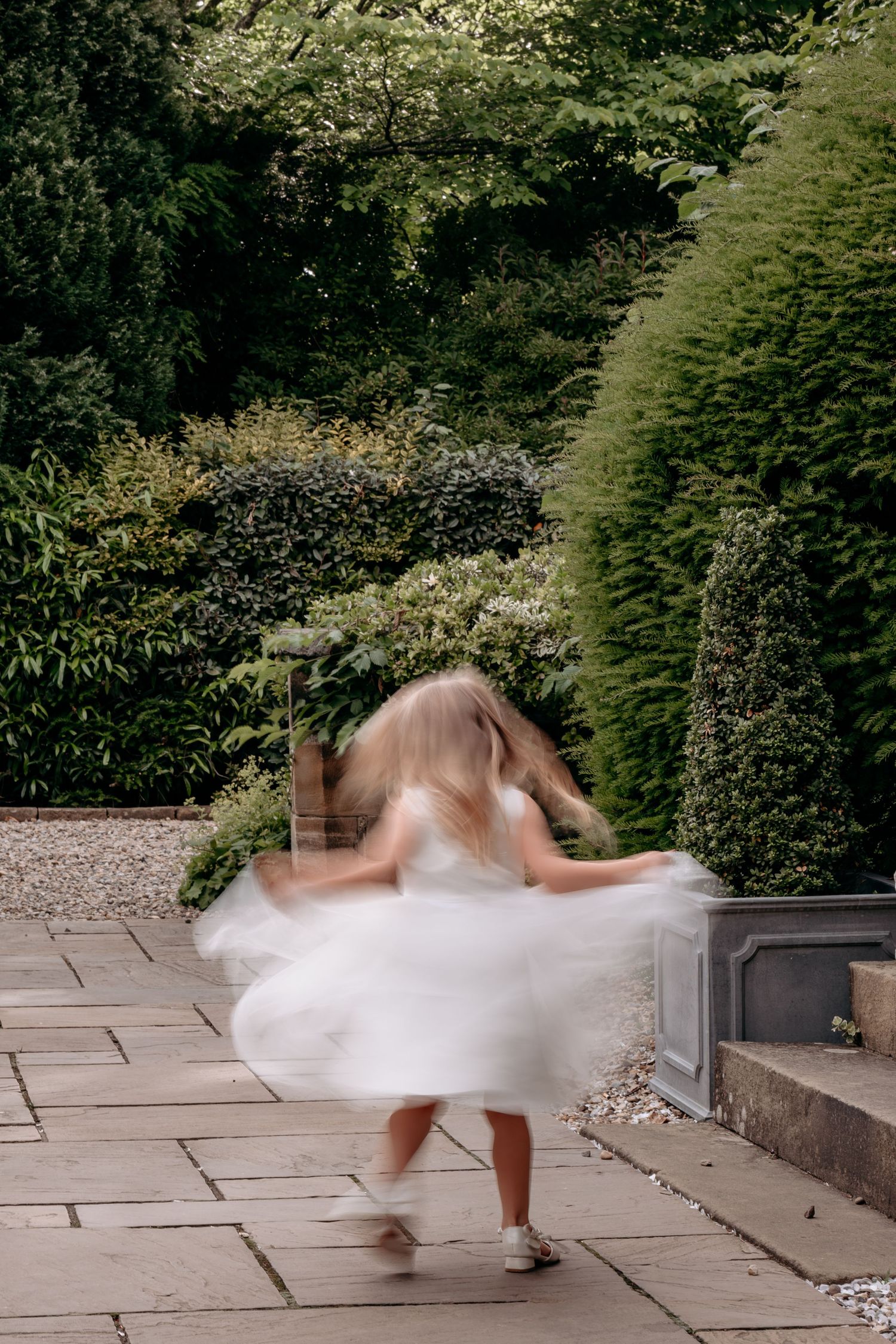Young girl in white dress spinning joyfully on stone patio surrounded by lush green garden hedges.