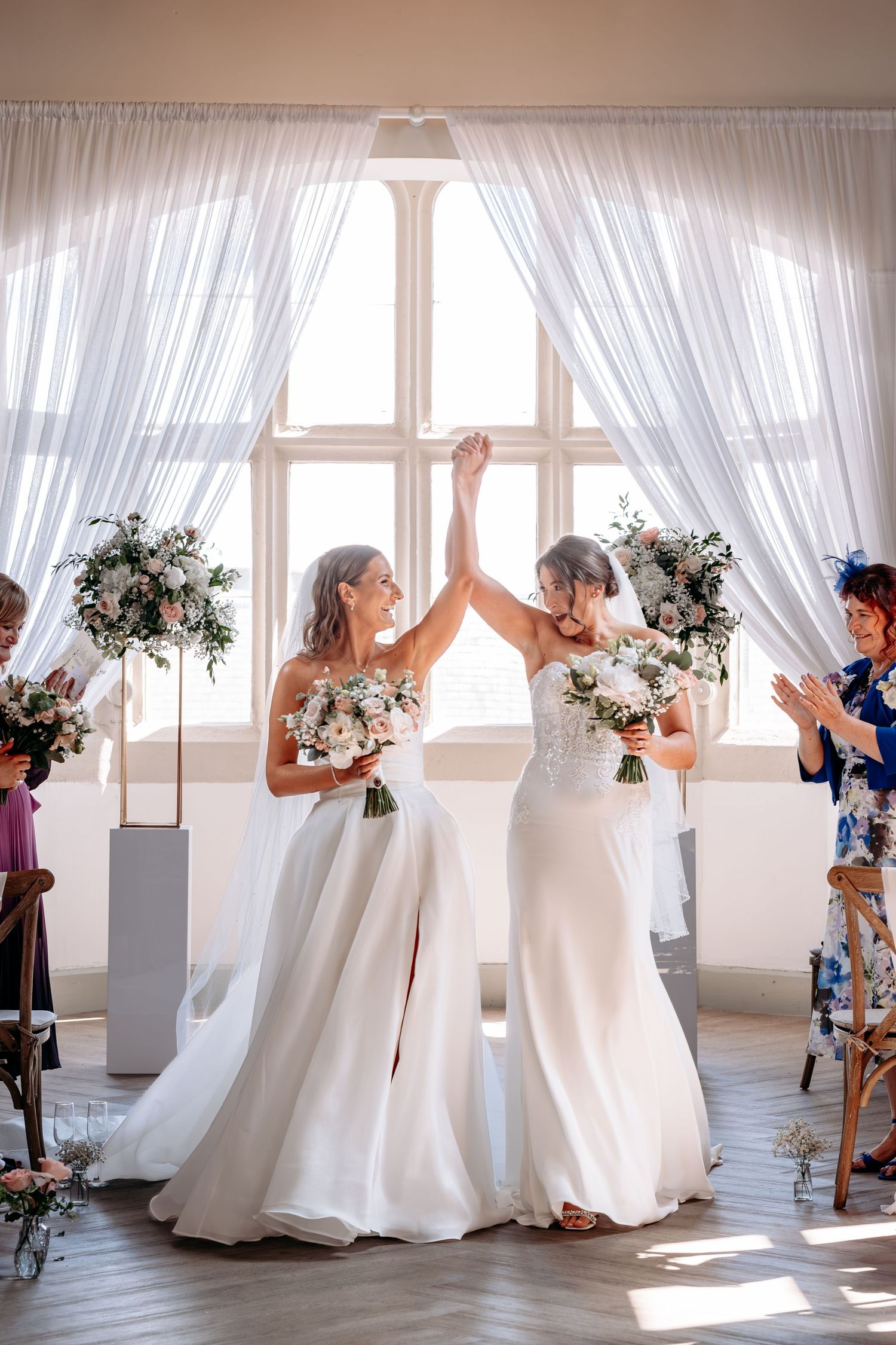 Two brides in white gowns celebrate their wedding ceremony, raising hands together while holding bouquets indoors.