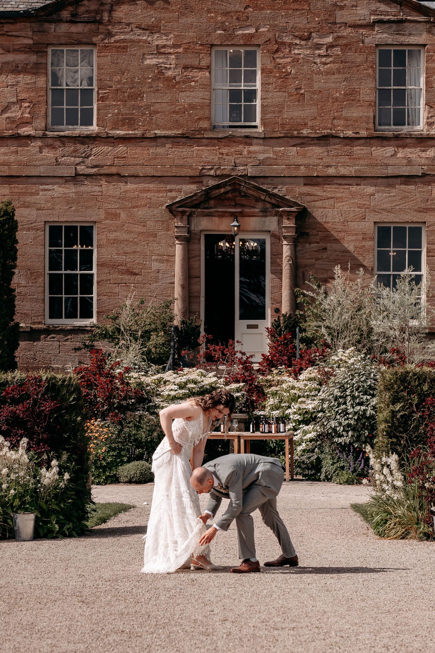 Bride and groom dancing in front of a grand red sandstone manor house surrounded by lush gardens.