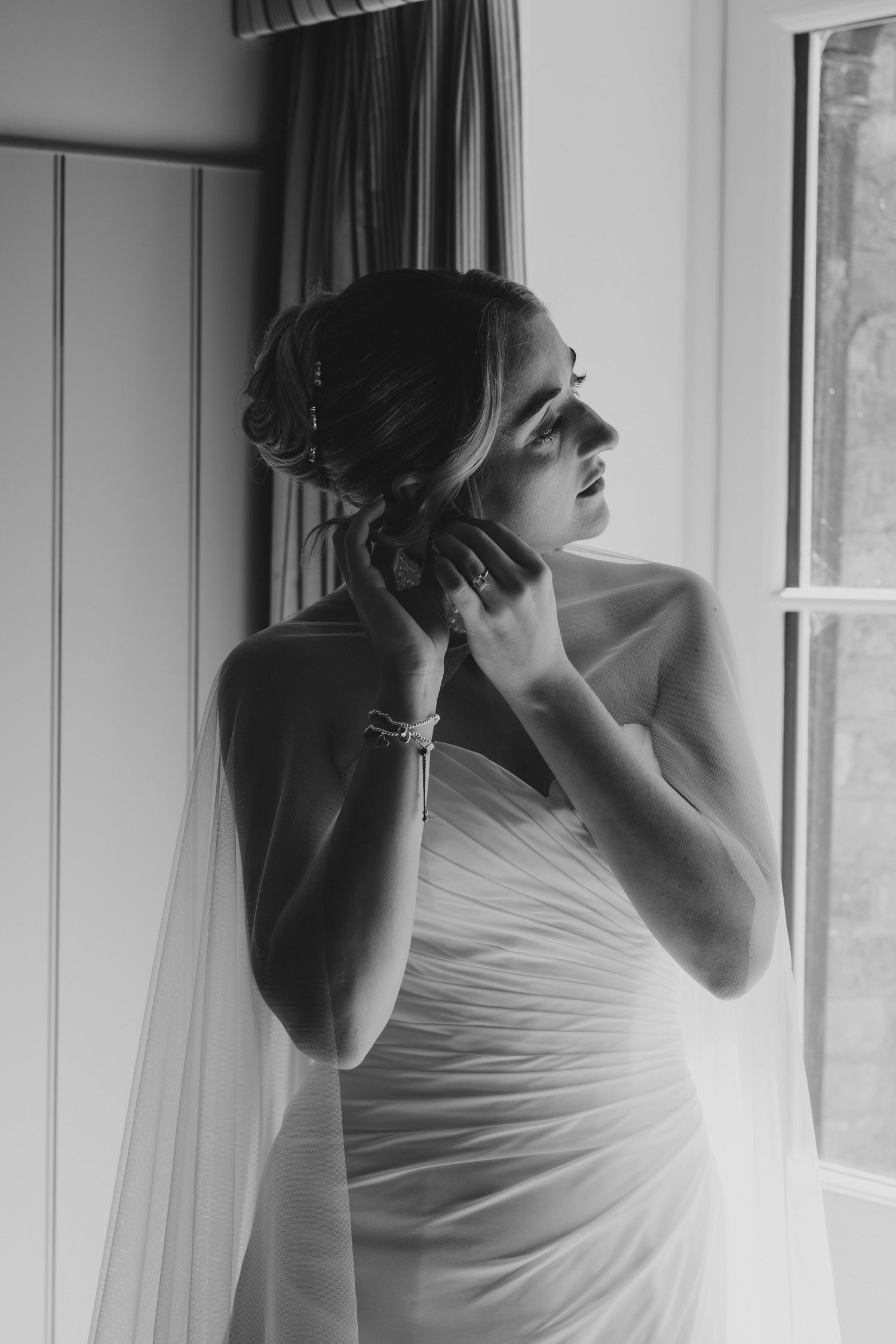 Black and white photo of a bride in a wedding dress adjusting her earring by a window on her wedding day.