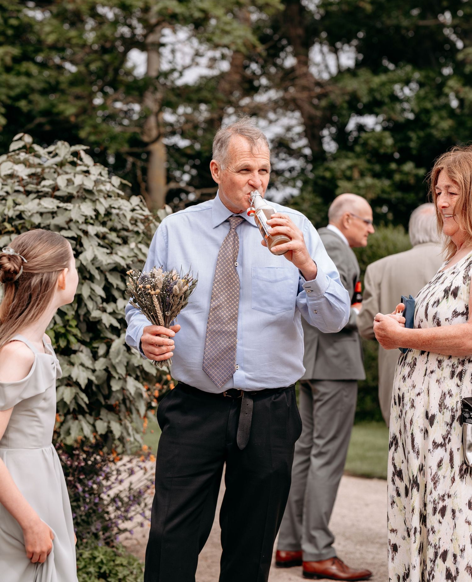 Man in blue shirt and tie eating at an outdoor wedding reception with guests socializing in a garden.
