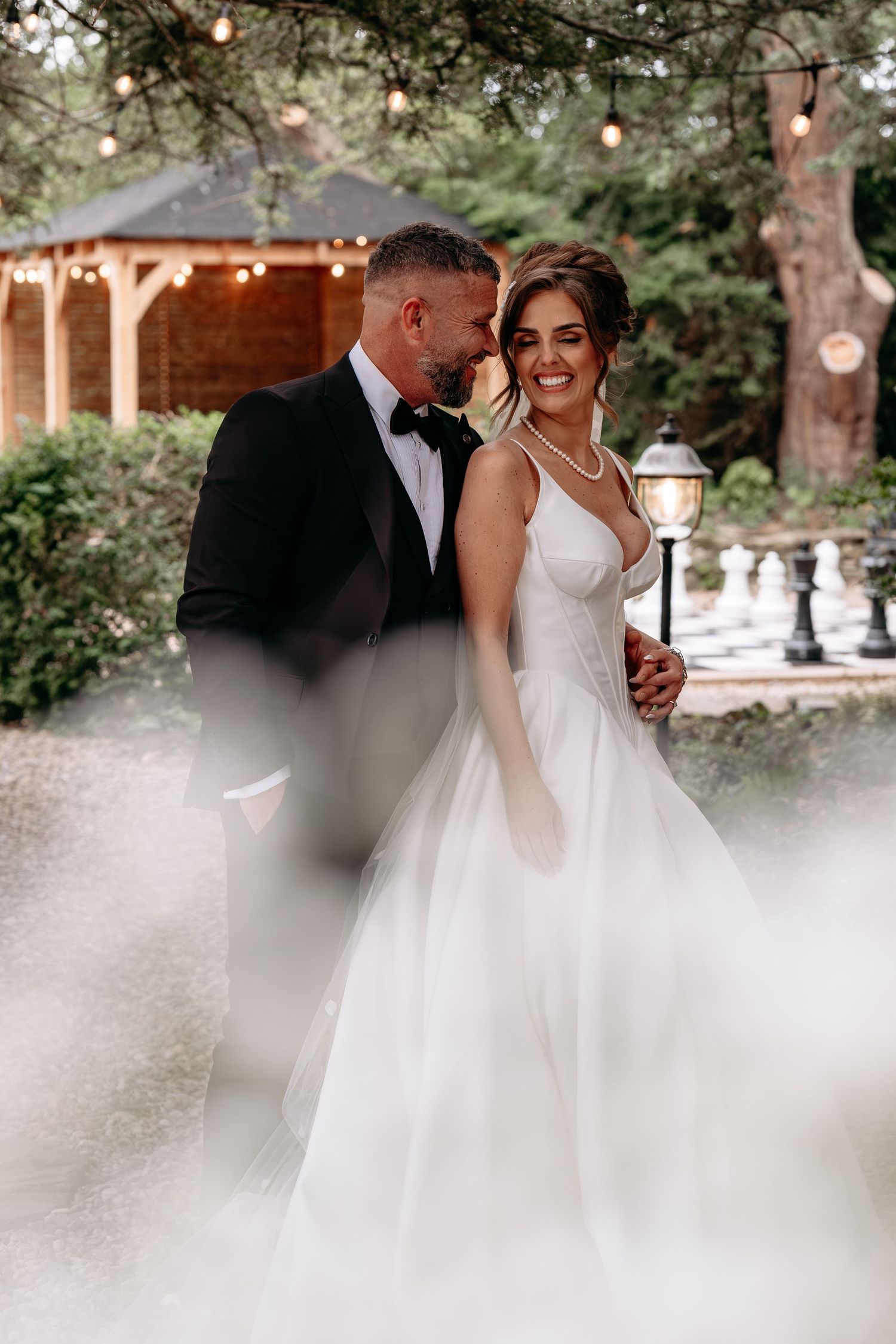 A bride in a white ballgown and groom in a black tuxedo share a romantic moment in a garden with string lights.