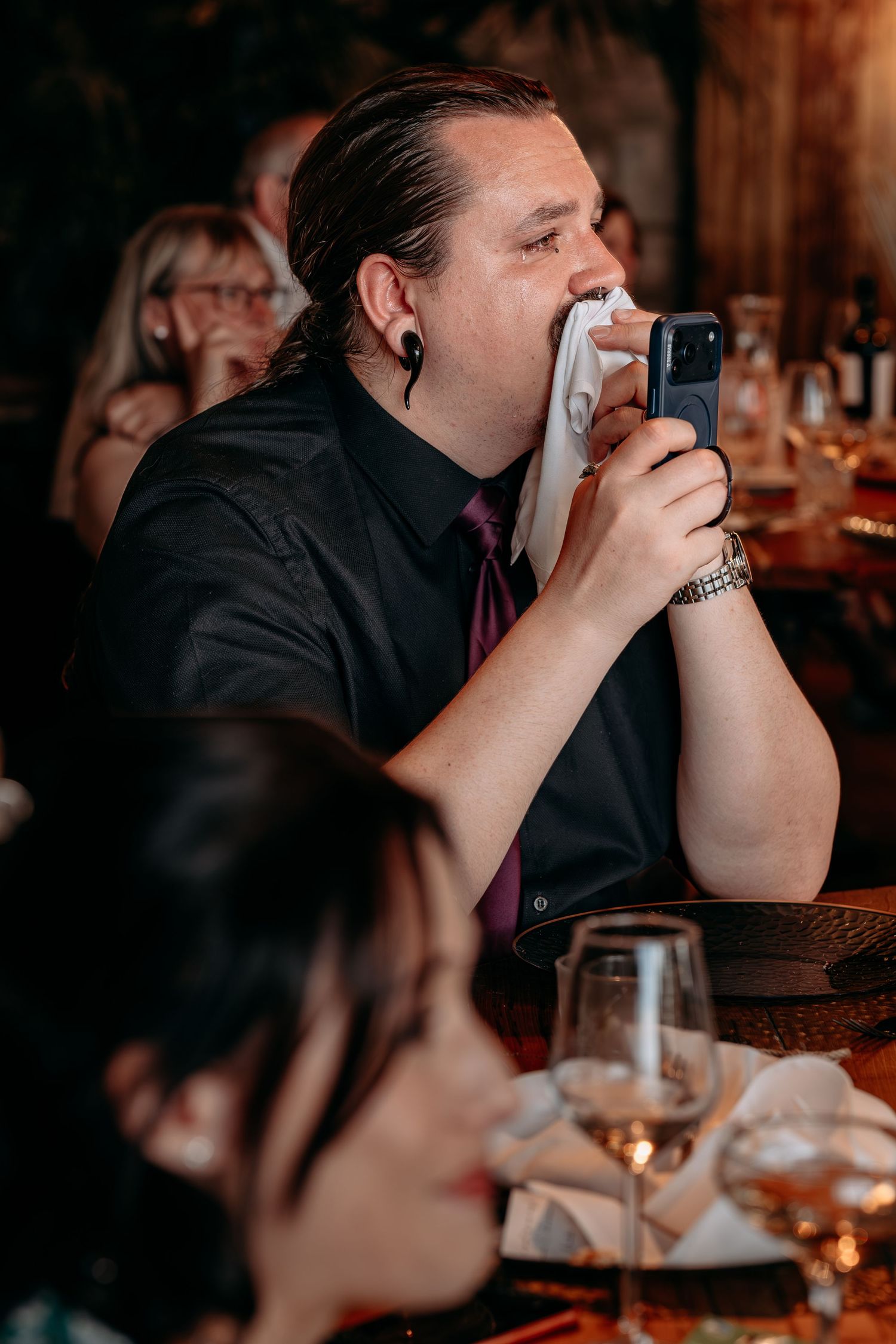 Emotional wedding guest wiping tears with napkin while holding phone at formal dinner reception.