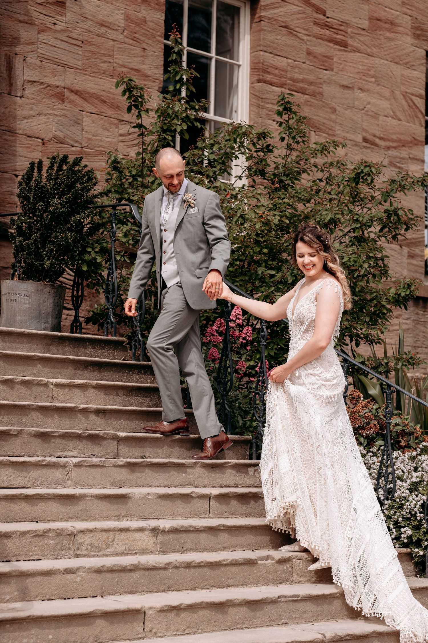 A bride and groom hold hands while descending stone steps outside a historic brick building on their wedding day.