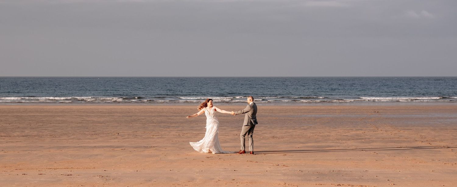 Bride and groom dancing on a sandy beach with calm ocean waves and cloudy sky in the background.