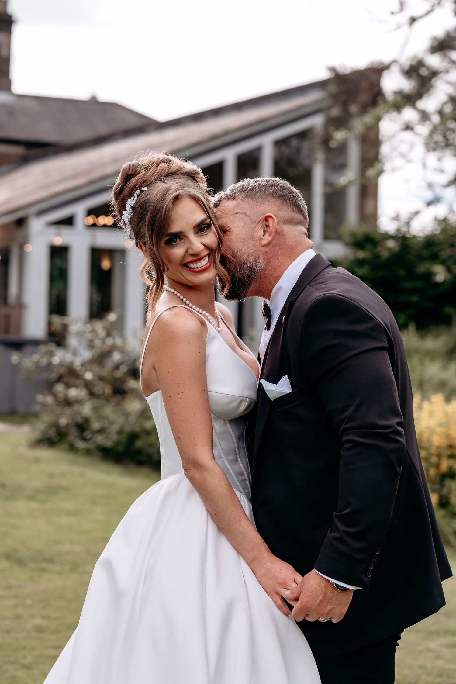 A smiling bride in a white gown receives a kiss on the cheek from her groom in a black tuxedo outdoors.