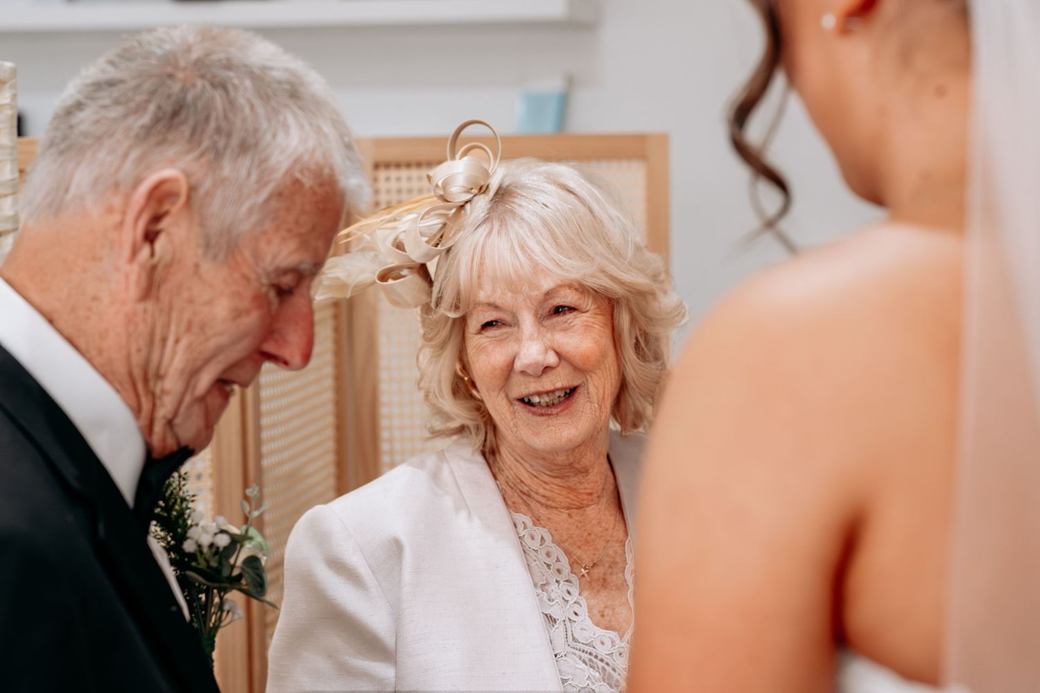 Elderly woman in fascinator hat smiling warmly at a wedding ceremony with bride and groom nearby.